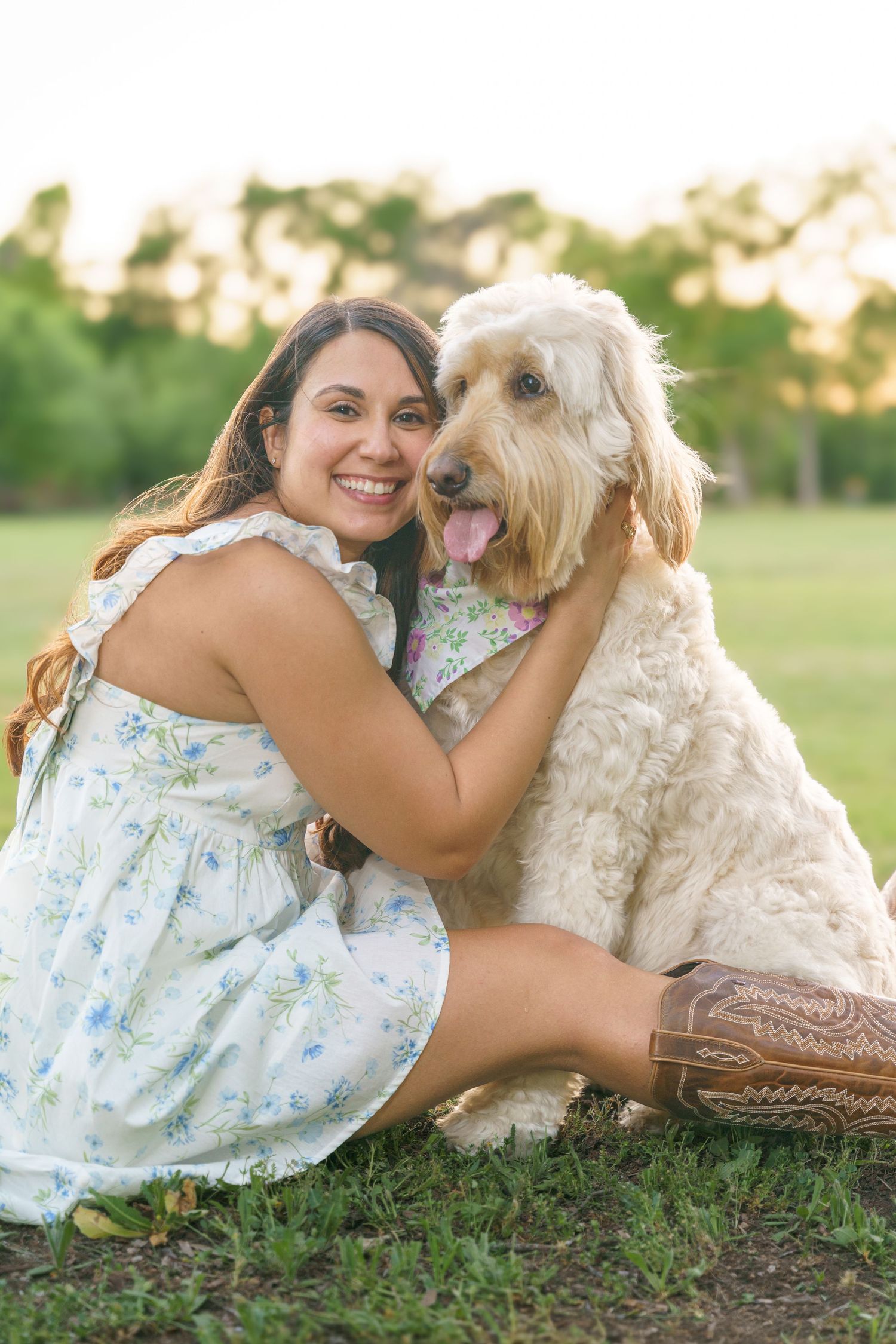 Woman in floral dress cuddling with goldendoodle dog on grassy field at sunset.