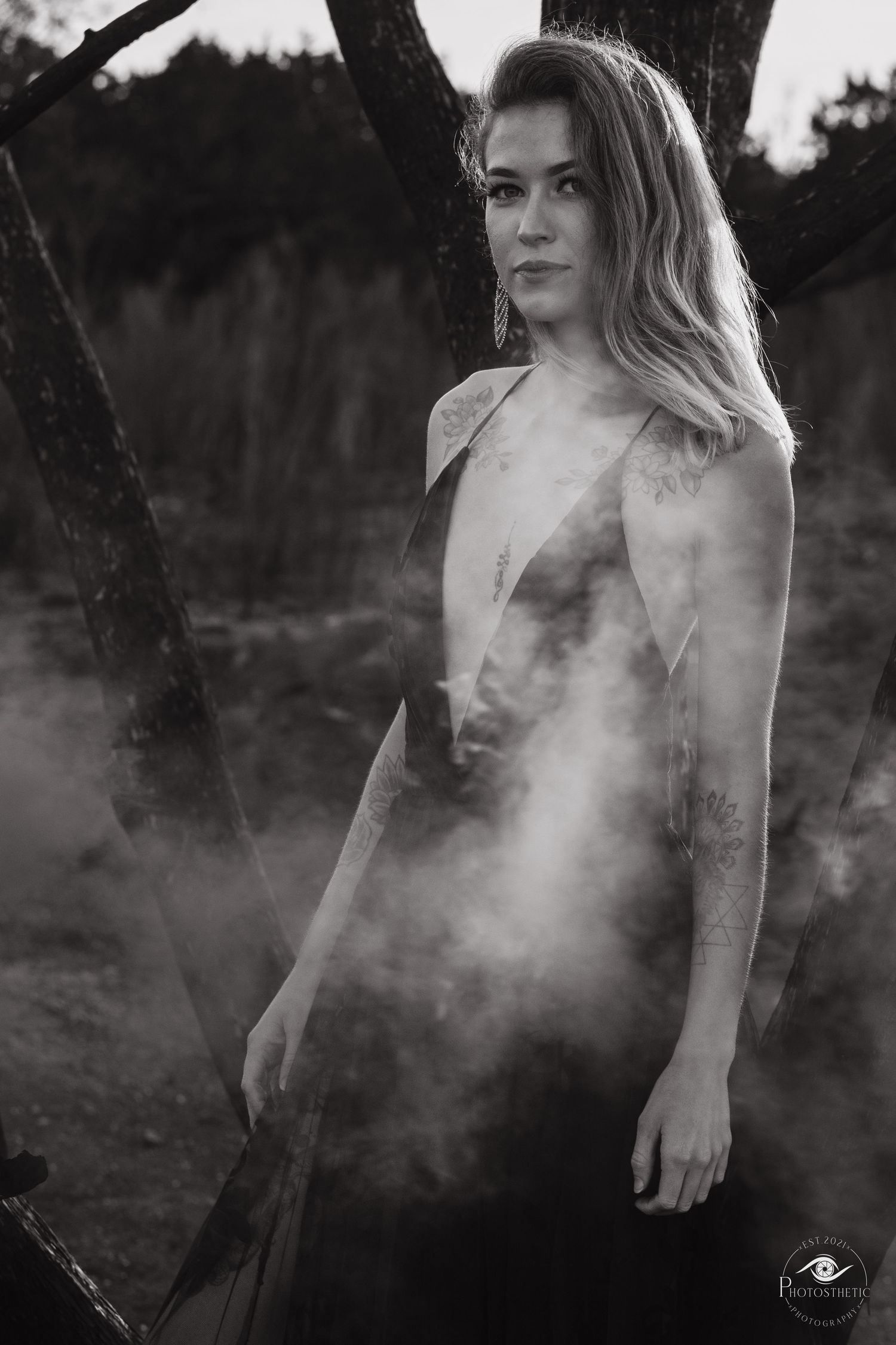 Artistic black and white outdoor portrait of a tattooed woman in an elegant dress with atmospheric smoke.