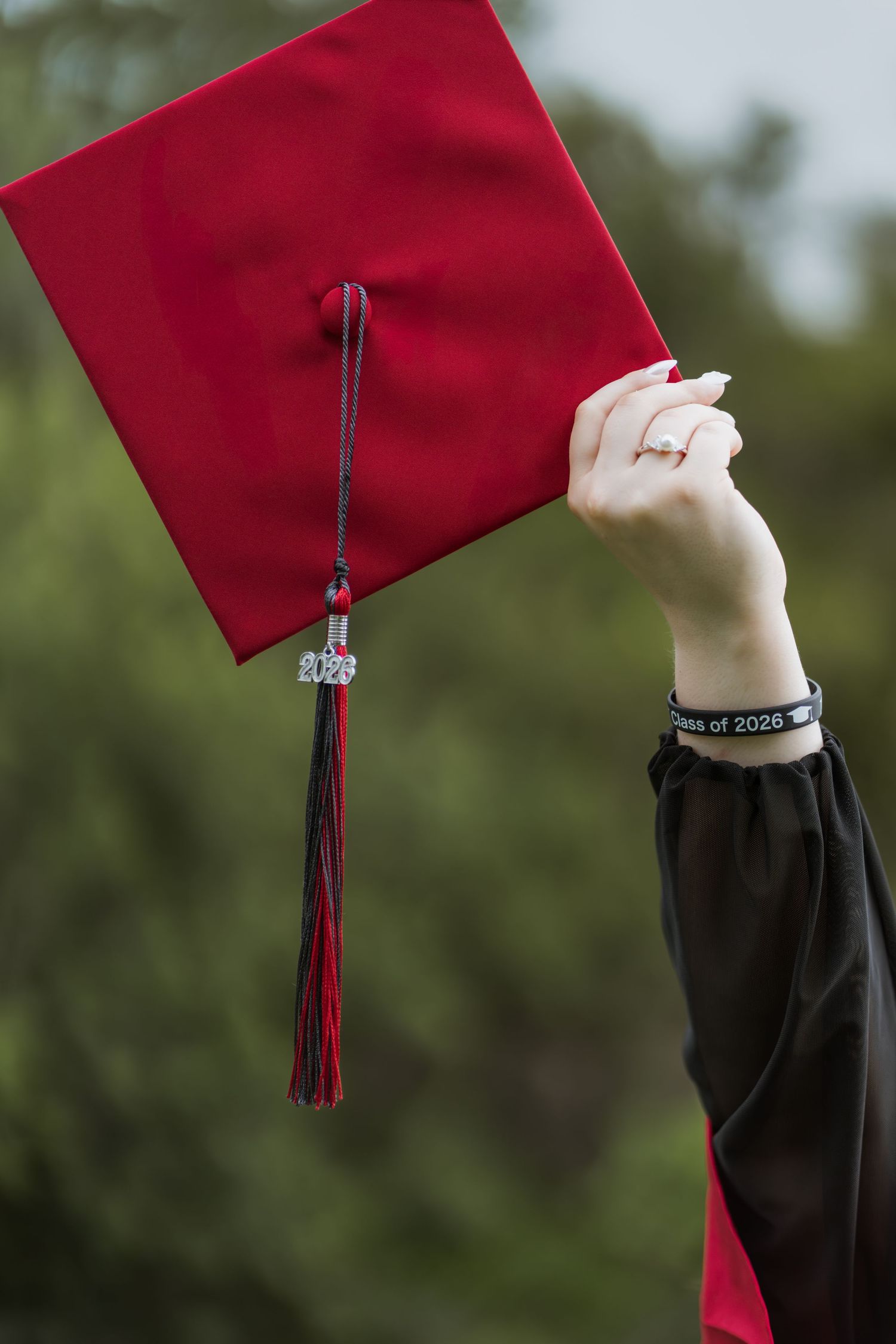 Hand holding a red graduation mortarboard cap with Class of 2026 tassel charm against green trees.