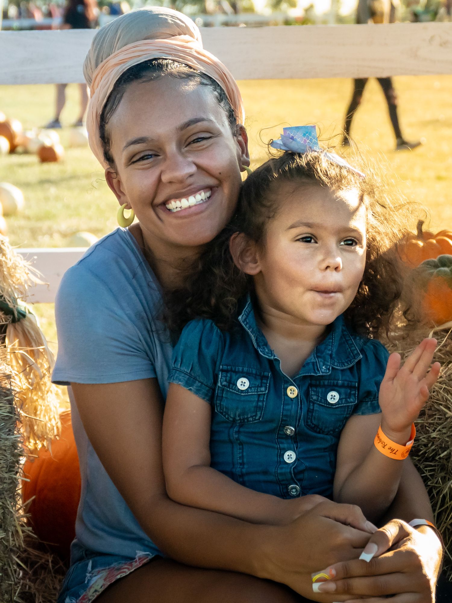 Mother and young child sitting on hay bales at a pumpkin patch, smiling during an outdoor fall portrait.