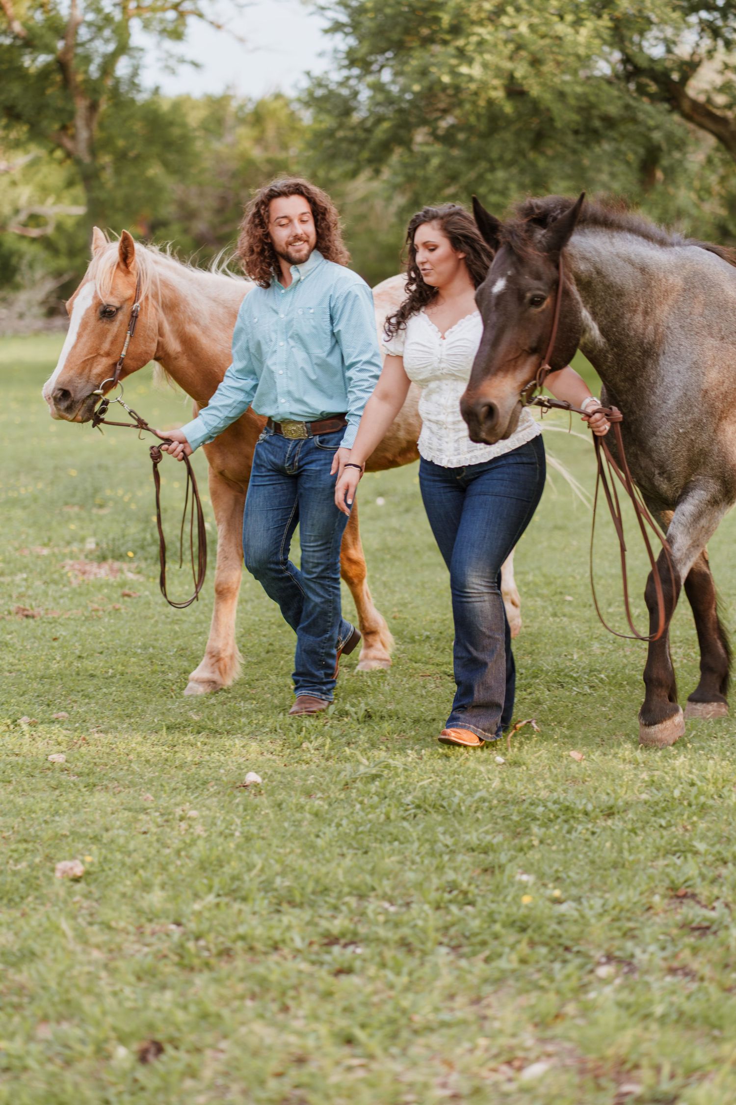 A couple walks two horses across a green field, wearing jeans and western shirts in an outdoor country setting.