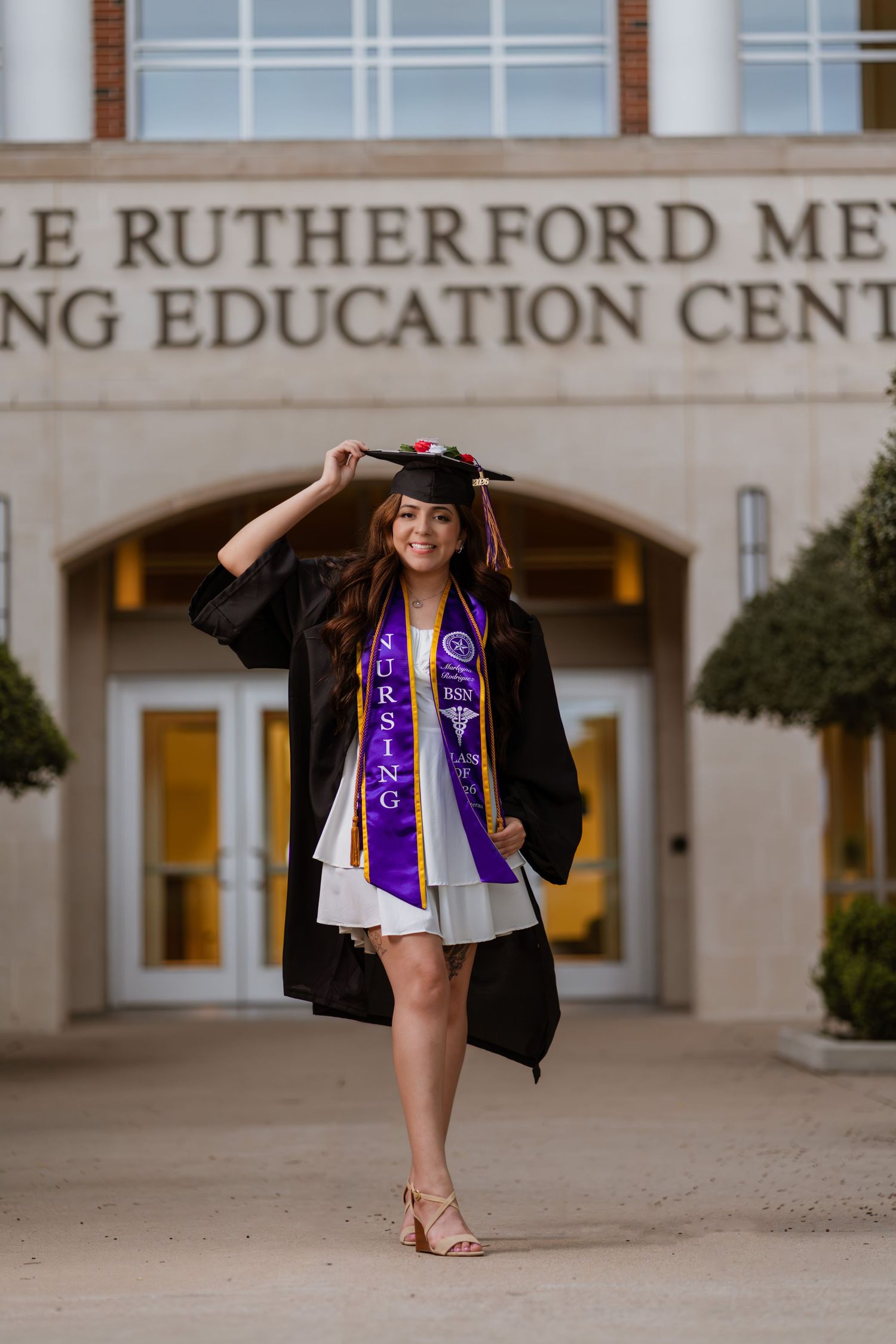 A nursing graduate poses in cap and gown with BSN stole outside a medical education center.