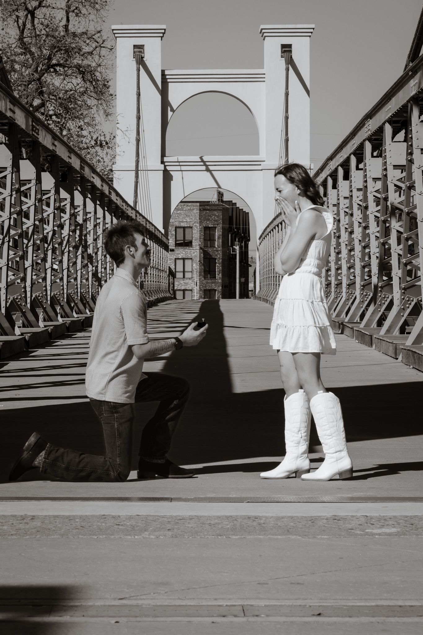 Man proposes on one knee to woman in white dress and boots on a bridge in black and white photo.
