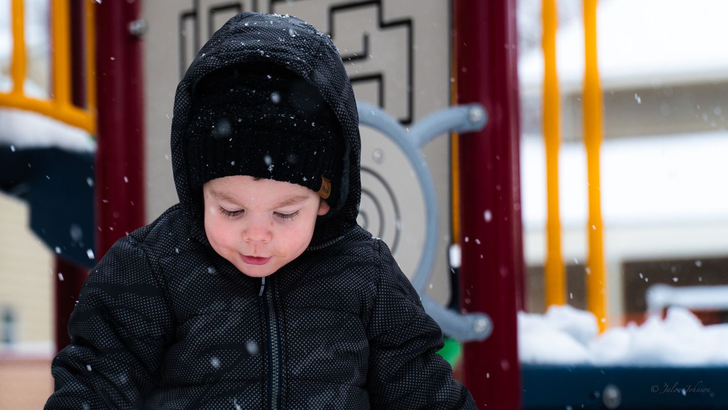 Toddler wearing a winter jacket and hat playing outdoors at a playground during a snowy day.