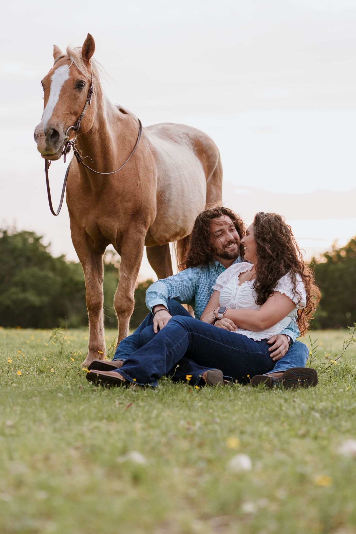 Couple sitting on grass smiling together with a palomino horse standing behind them in a green field.