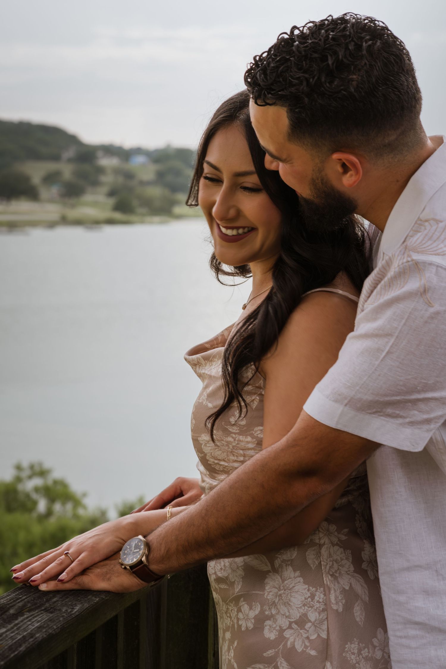 Couple sharing a romantic moment on a wooden railing overlooking a lake, woman smiling wearing floral dress.