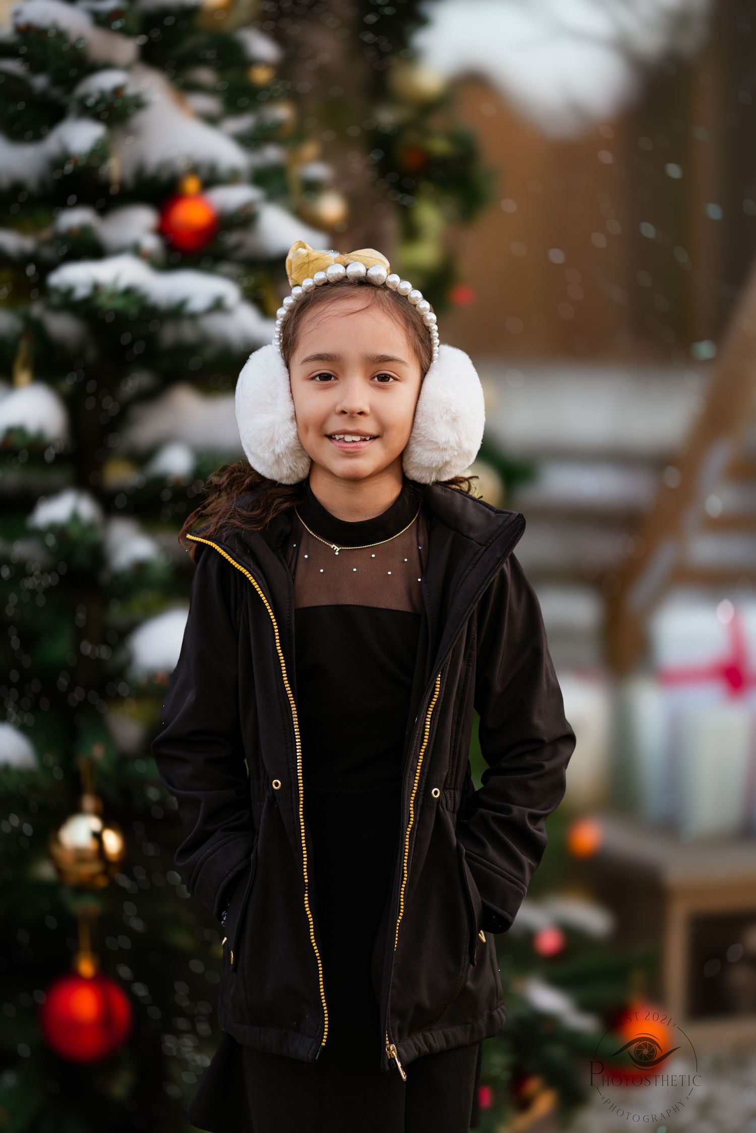 Winter portrait of a person in black coat and white earmuffs standing by a snow-covered Christmas tree with red ornaments.