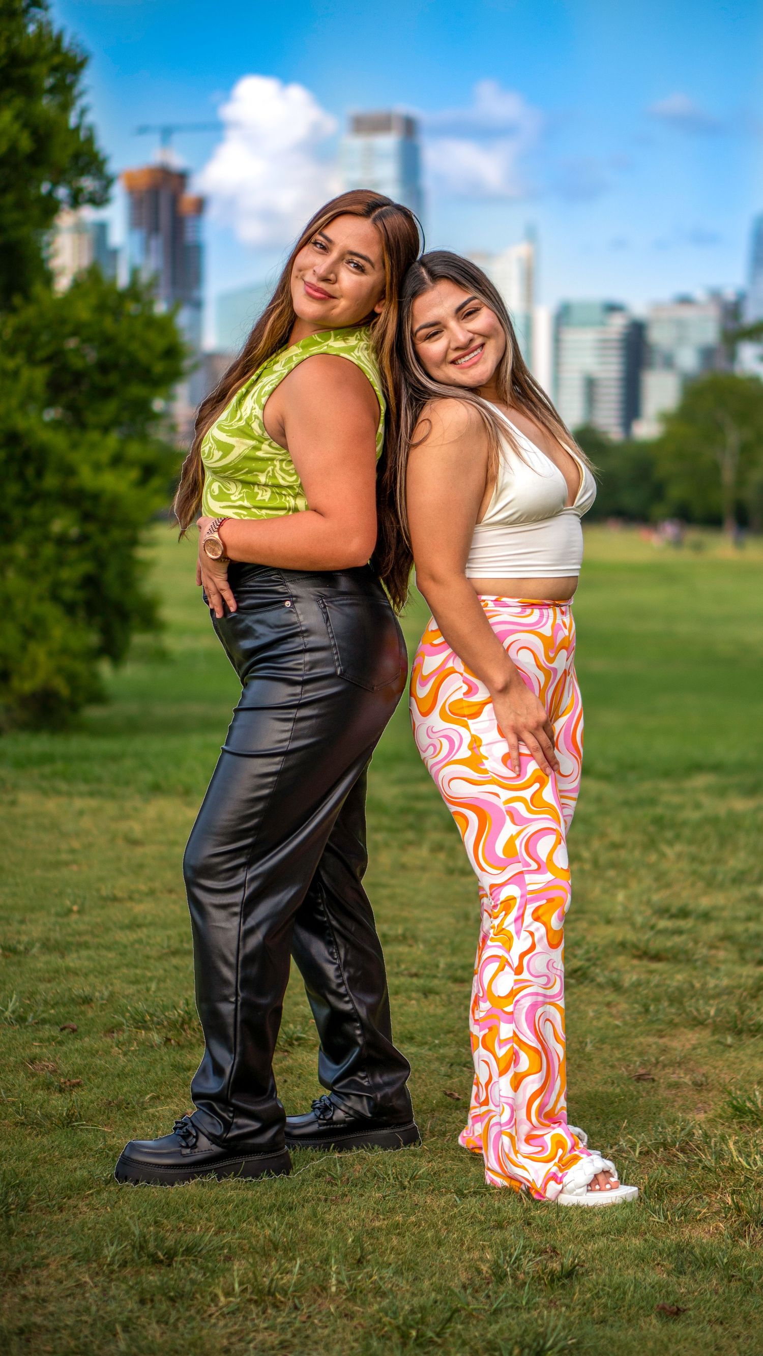 Two women standing back-to-back smiling in a park with a city skyline in the background.