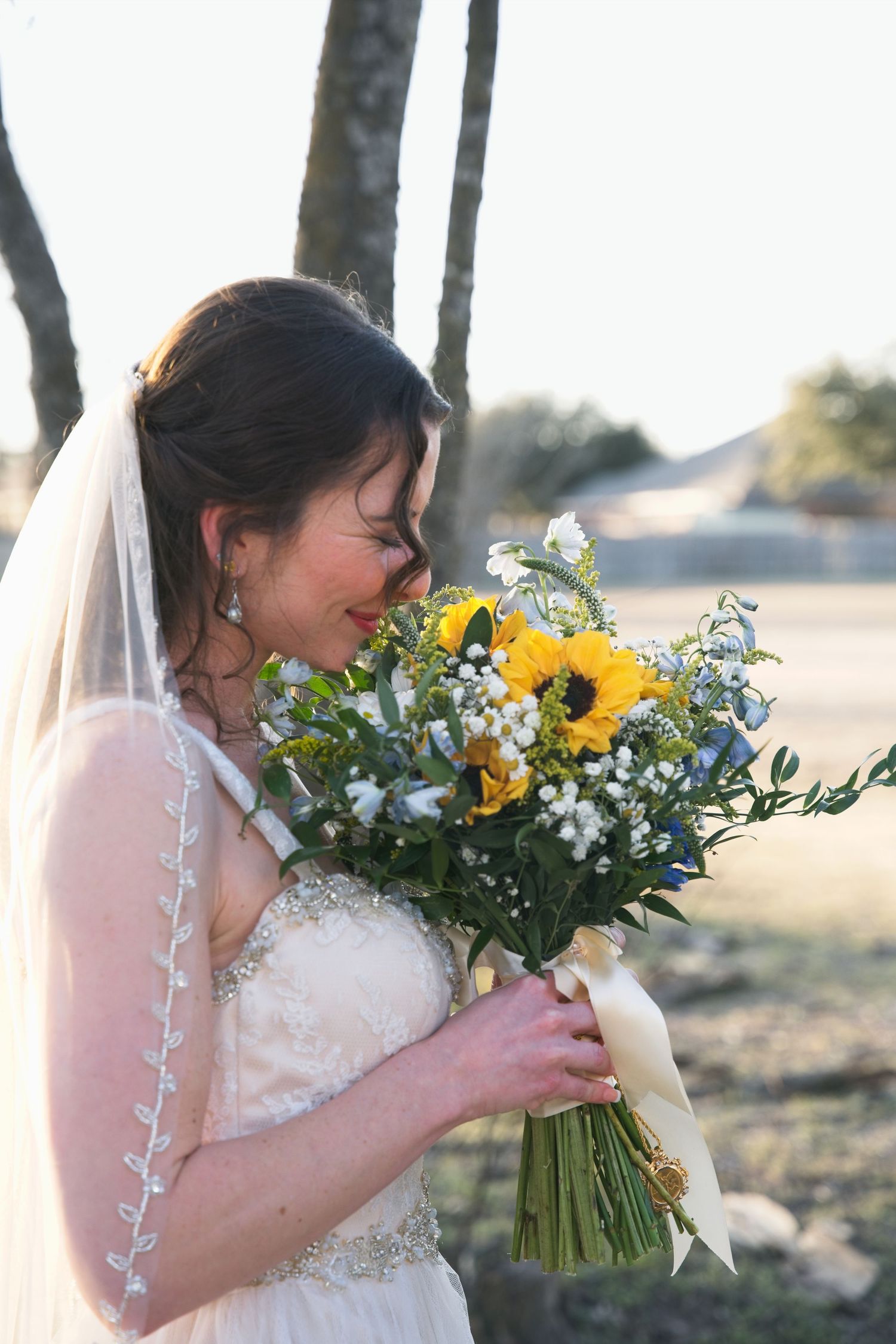 A bride in a white lace dress smells a vibrant bouquet of sunflowers and mixed florals against an outdoor backdrop.