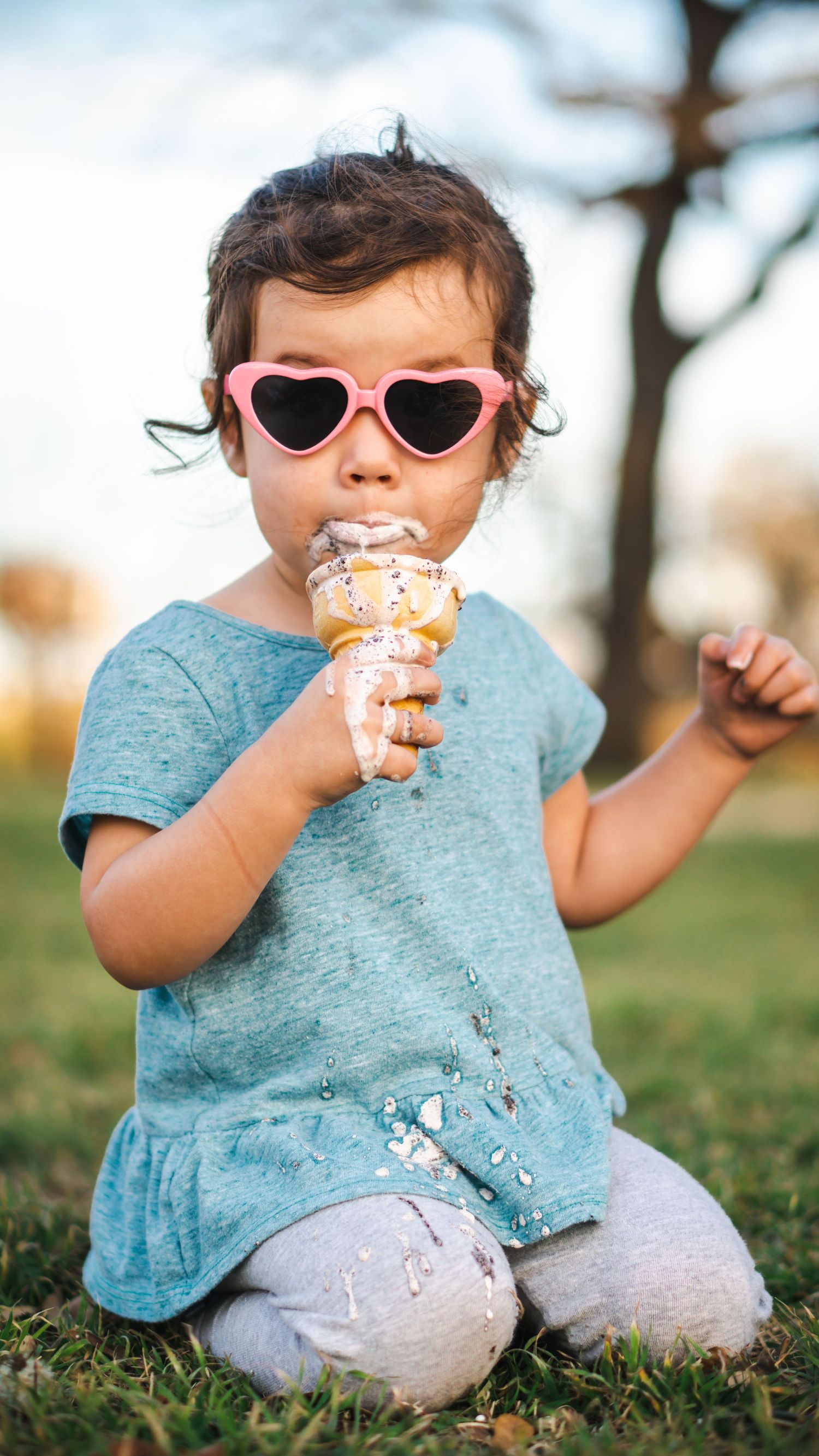 Young child wearing heart-shaped sunglasses eating ice cream outdoors in a park.