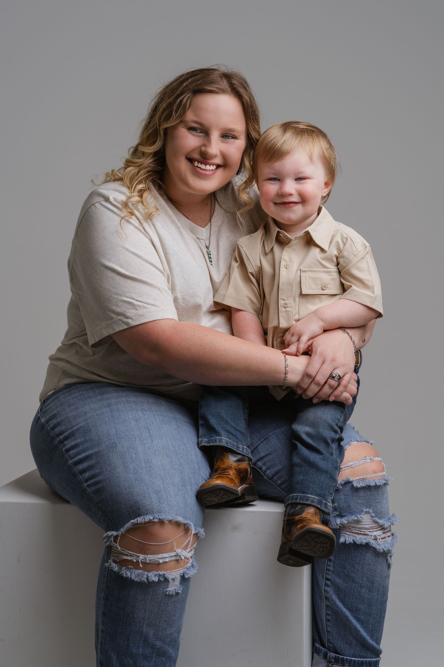 A smiling woman and toddler boy in matching beige shirts and jeans pose together for a studio portrait.