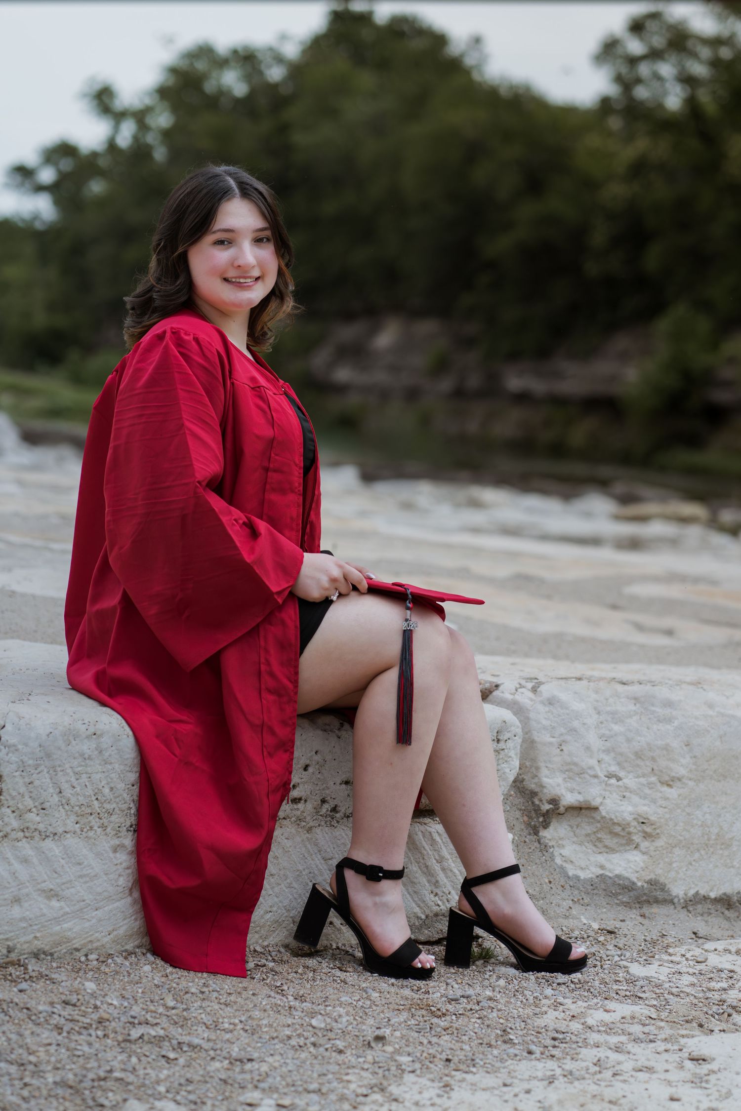 Young woman in red graduation gown and heels sitting on rocks outdoors, smiling and holding her cap.