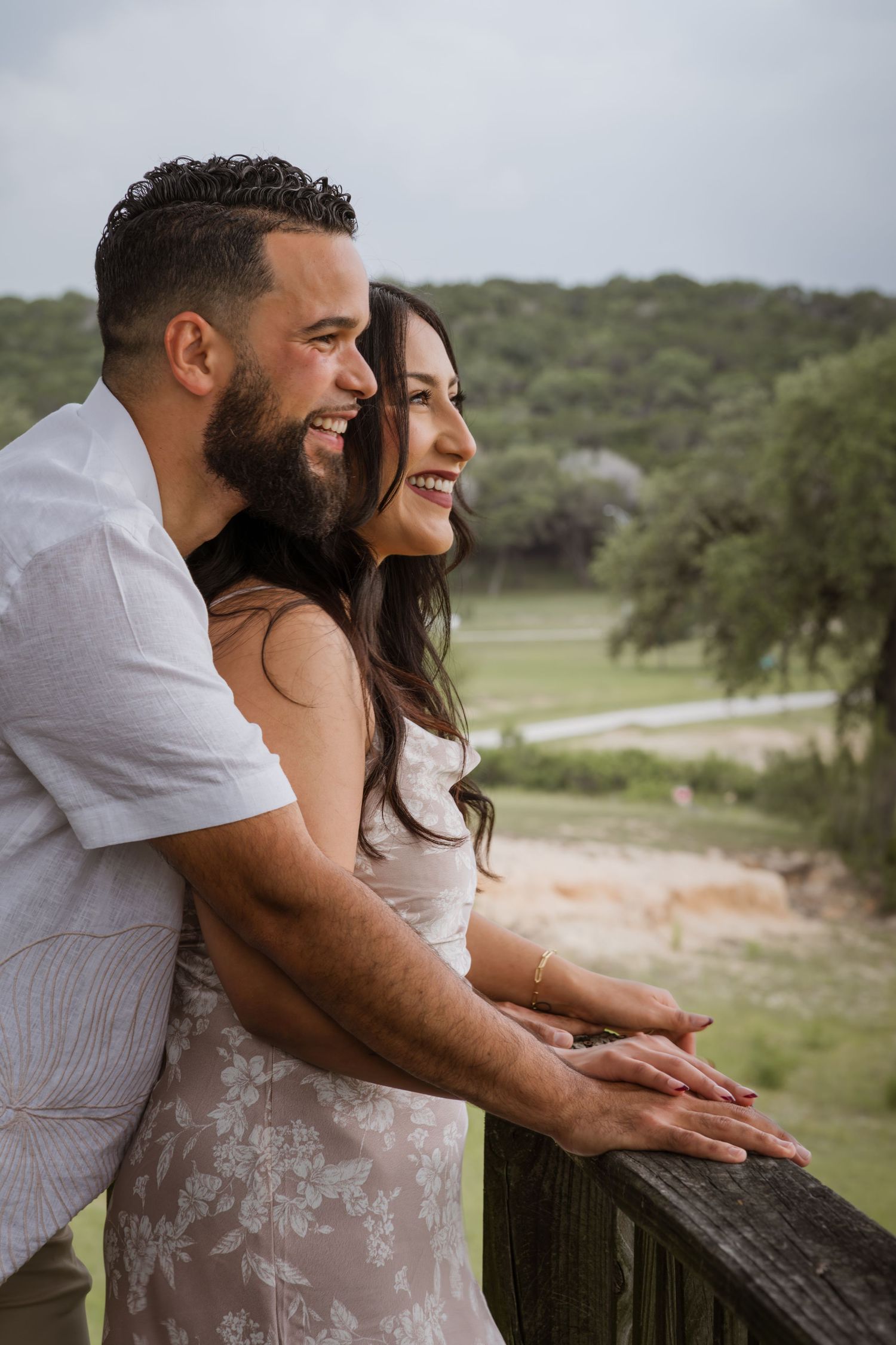 A smiling couple leans on a wooden railing, gazing at a lush green landscape on a cloudy day.