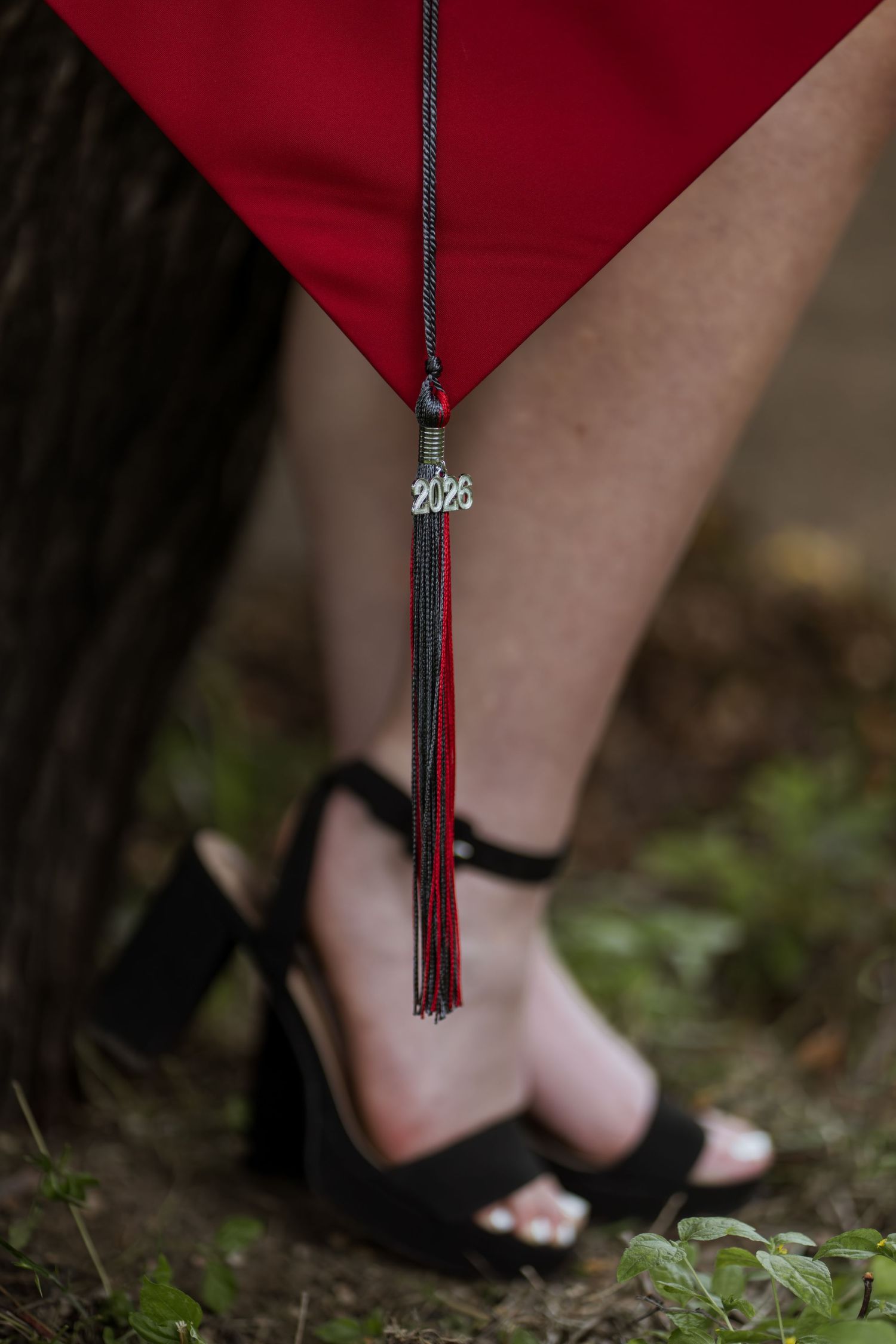 Close-up of graduation cap tassel with 2026 charm, red gown, and black heeled sandals on grass.