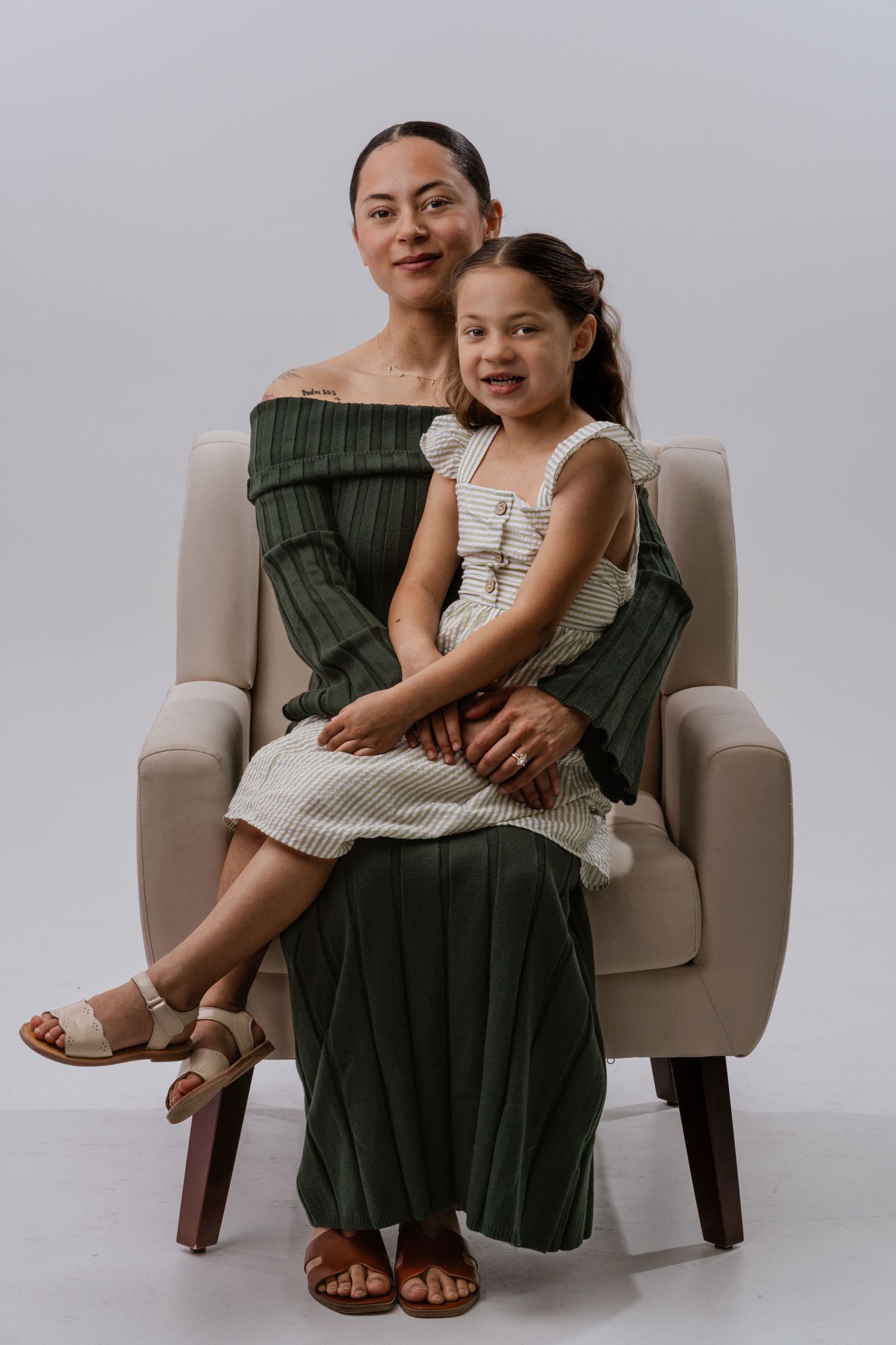 Woman in green dress sitting in armchair with young girl in striped dress on her lap, studio portrait.