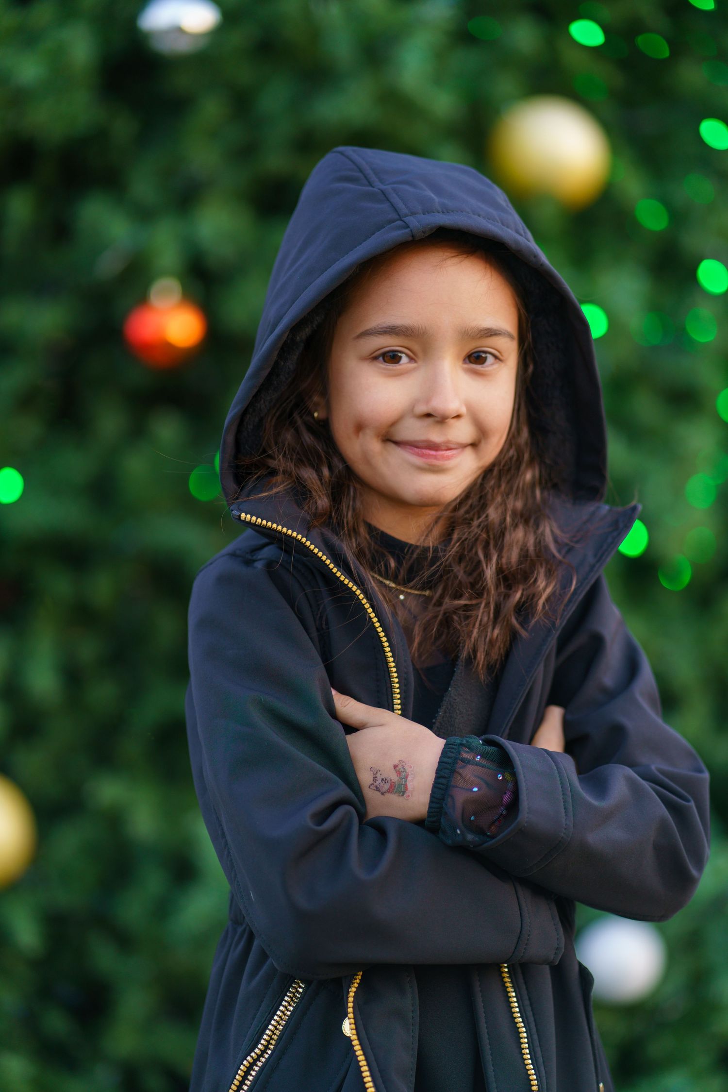 Young girl in black hooded jacket with arms crossed, smiling in front of a decorated Christmas tree.