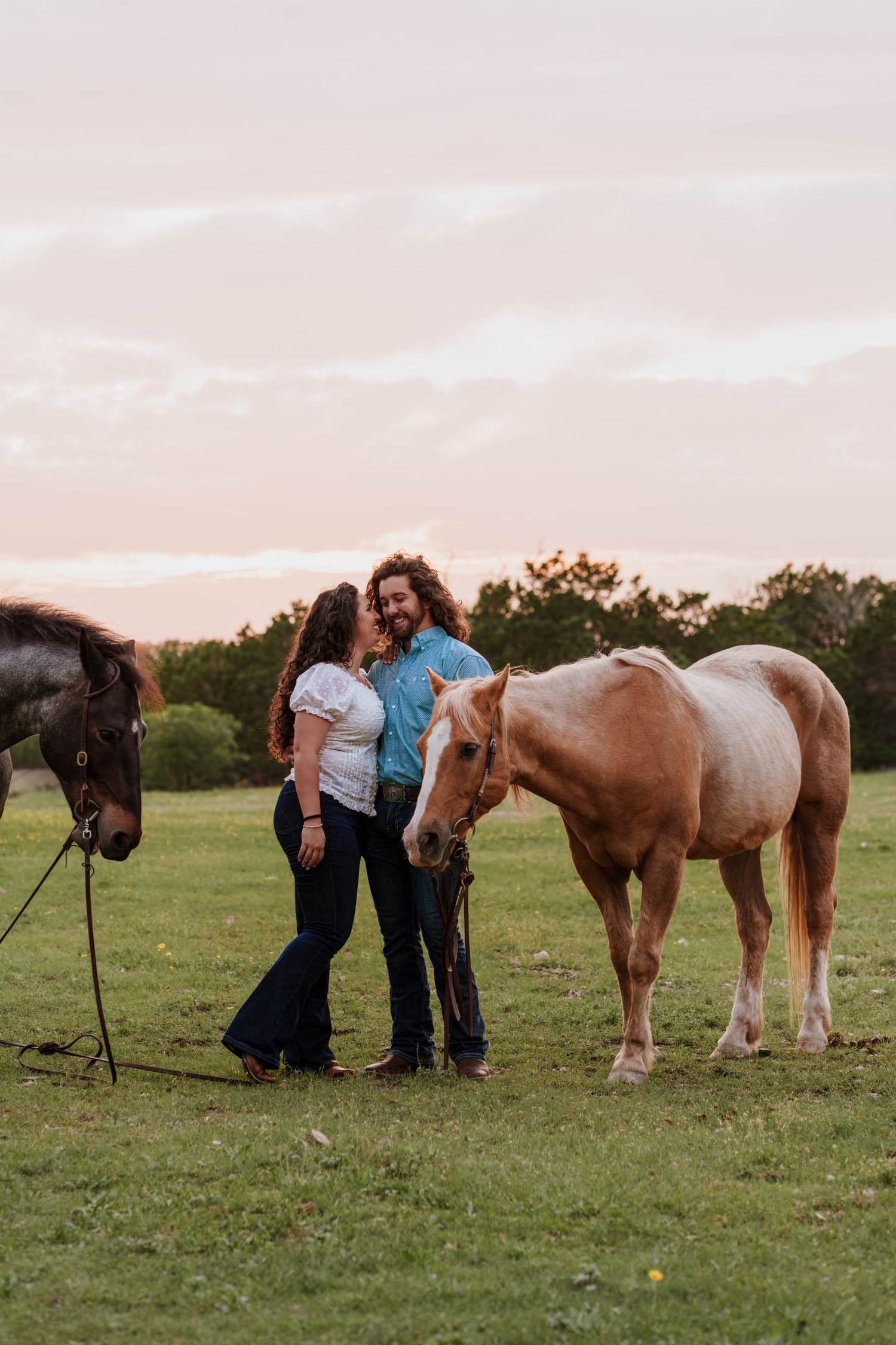 Couple sharing a romantic moment between two horses at golden hour on a green pasture.
