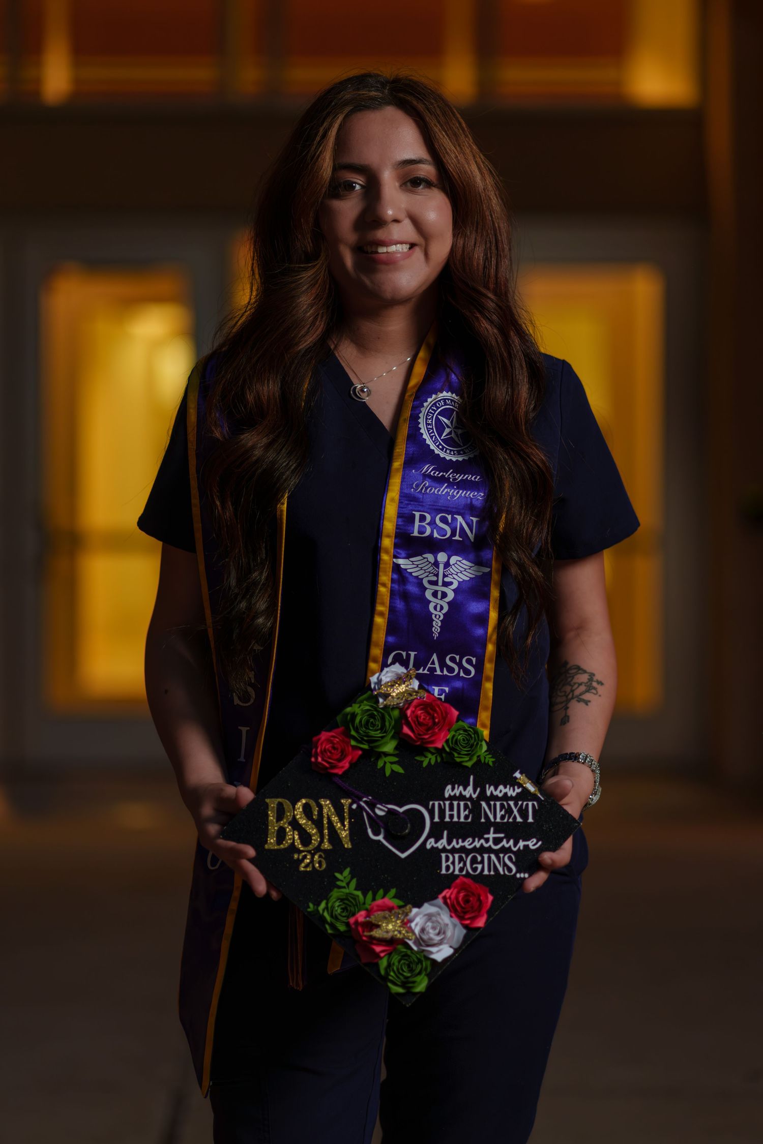BSN graduate smiling in navy scrubs holding decorated graduation cap with flowers and 'The Next Adventure Begins' quote.