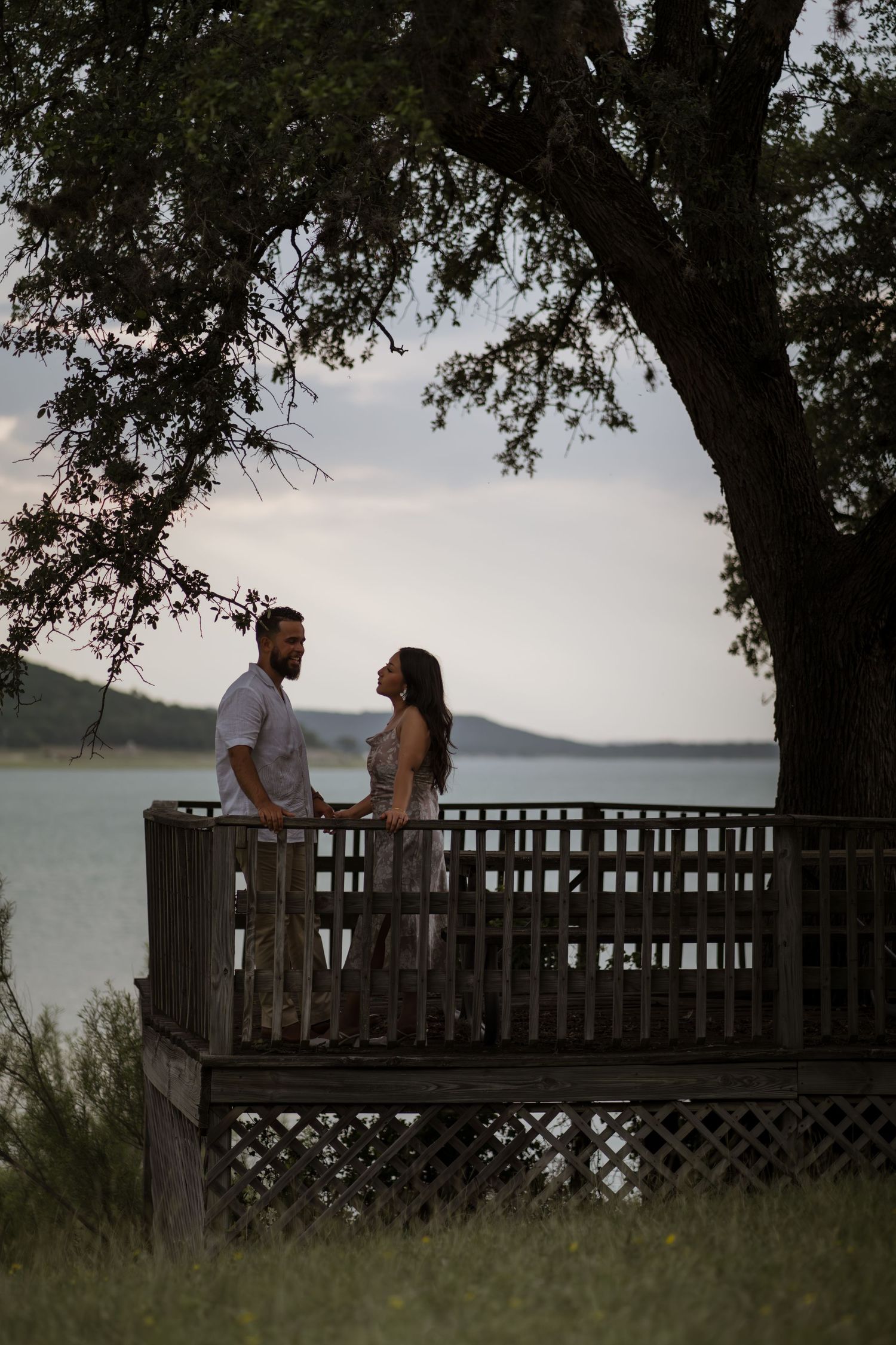 Romantic couple holding hands on a wooden deck by a lake at dusk, framed by a large tree.