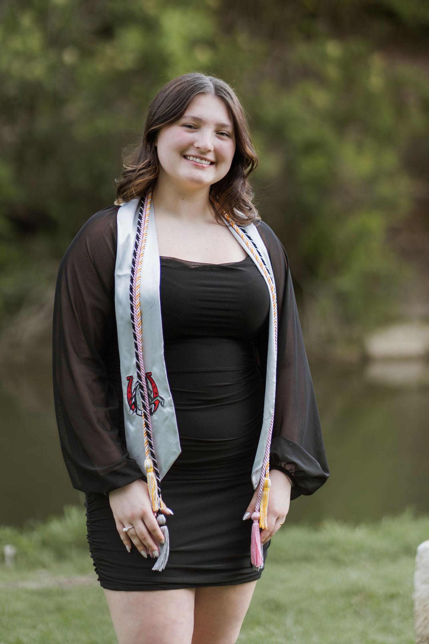 Smiling graduate in black dress wearing colorful honor stoles poses outdoors near greenery for graduation photos.