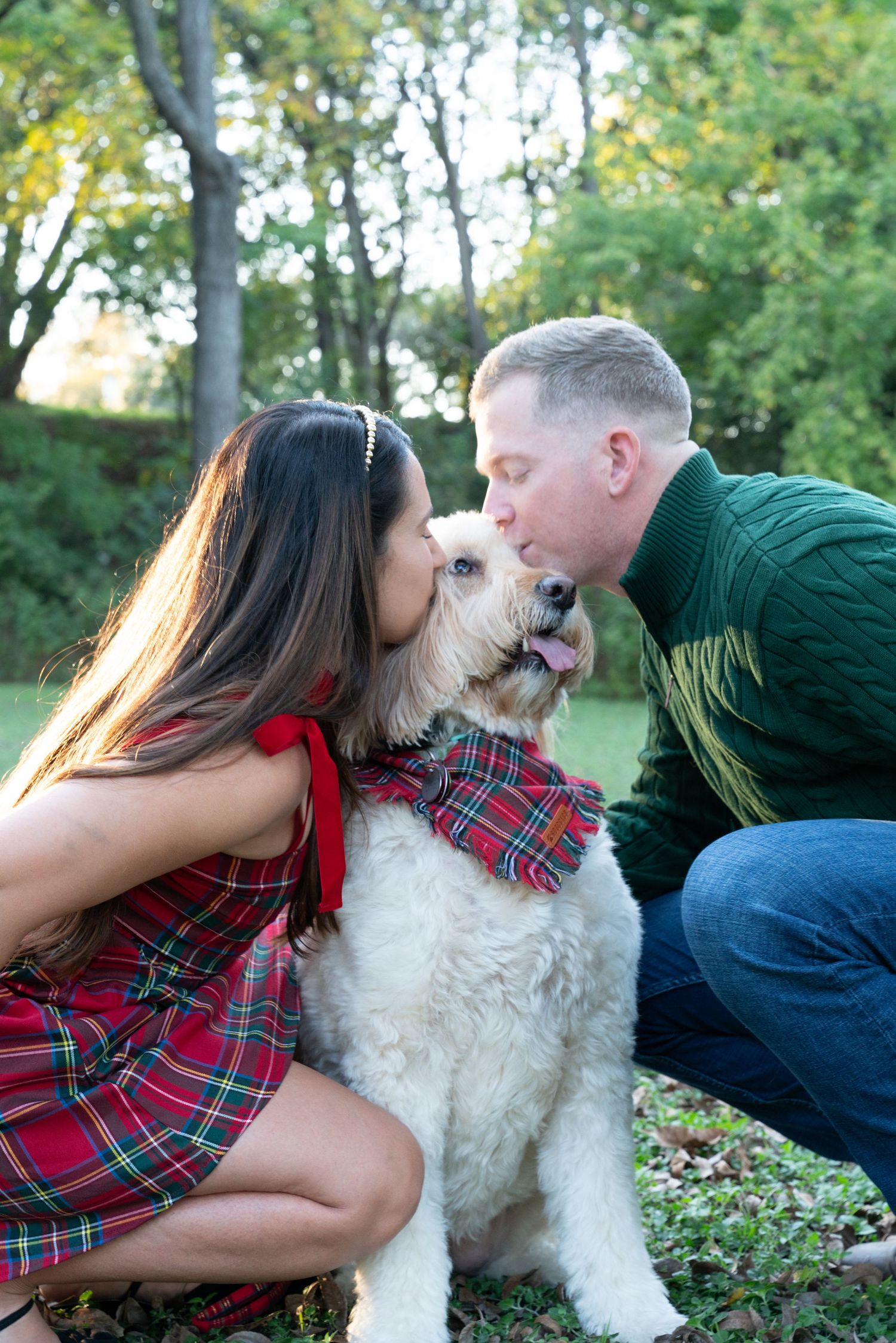 Couple in plaid outfits kissing their white Goldendoodle dog wearing a tartan bandana outdoors.