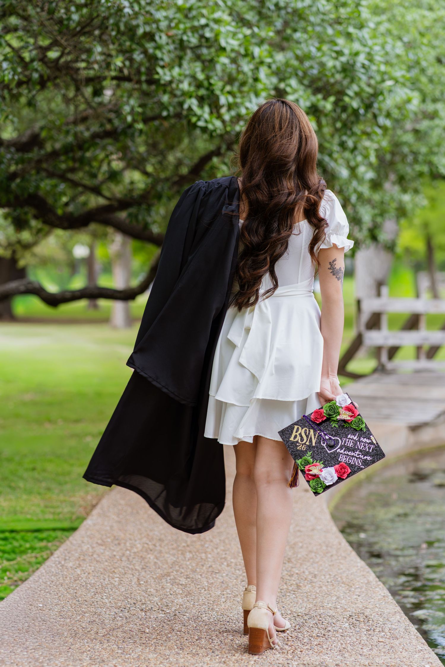 A nursing graduate in a white dress holds a decorated BSN cap and black gown while walking along a garden path.