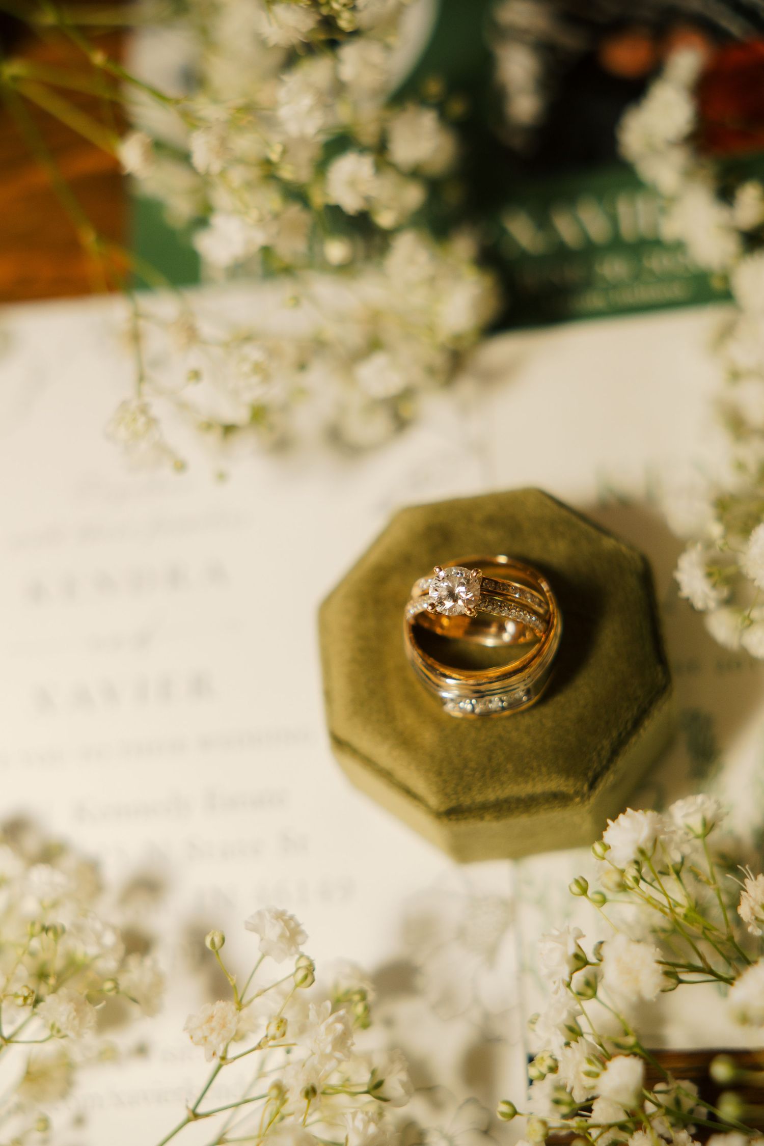 Gold wedding rings with diamonds displayed on a green velvet ring box, surrounded by baby's breath flowers and wedding invitations.