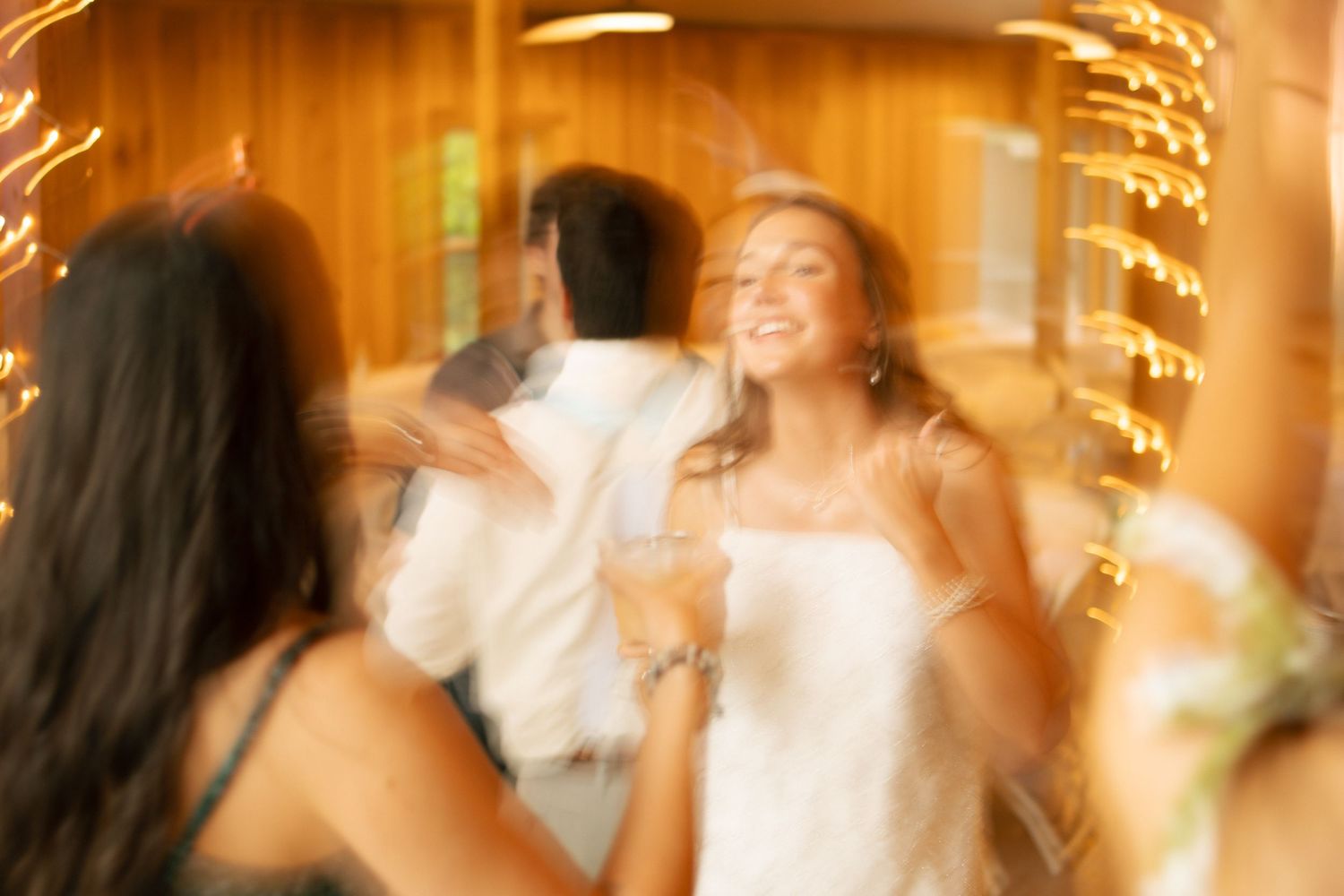 Blurred motion photo of bride laughing and dancing with guests under warm golden lights at wedding reception.