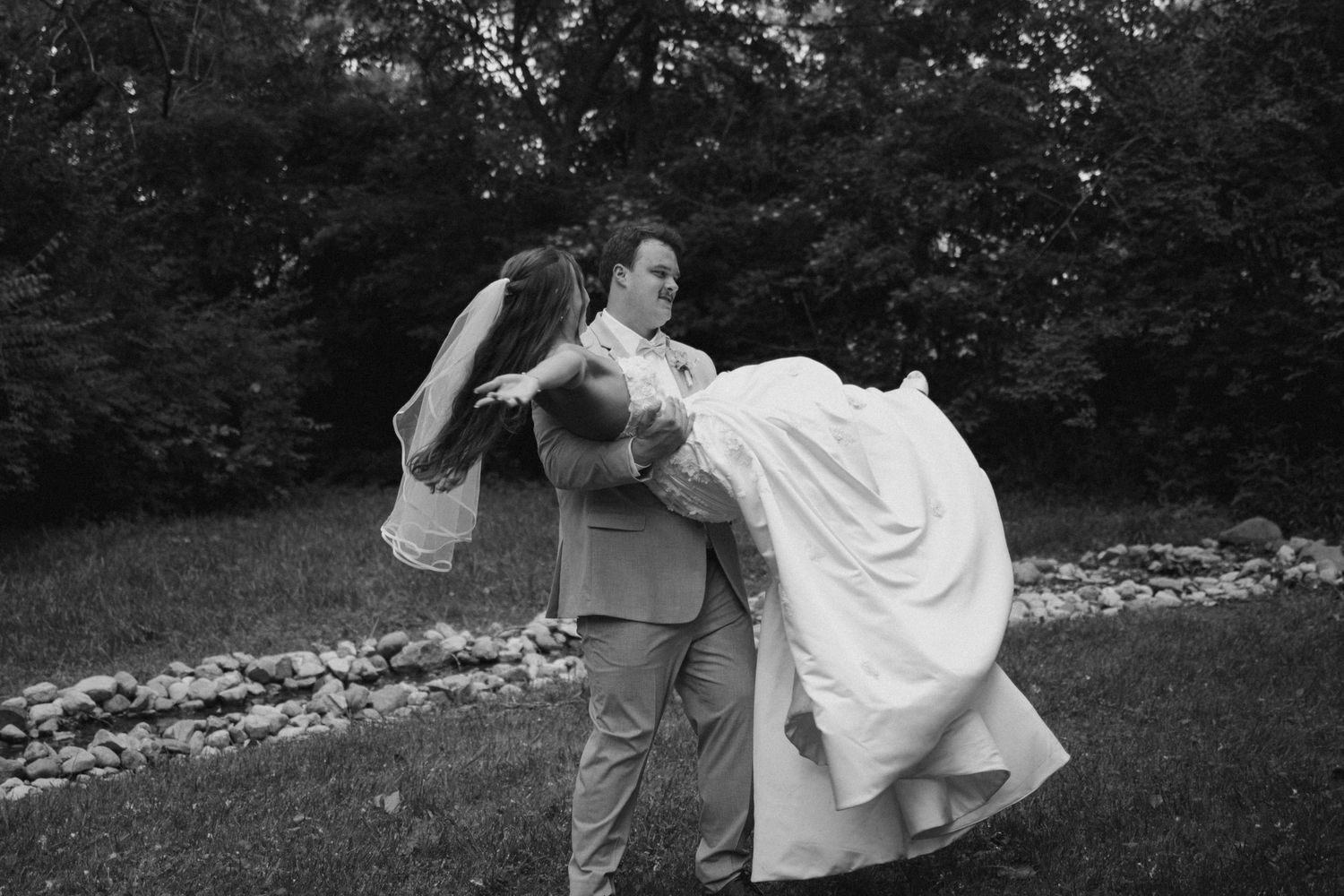 Groom lifting bride in wedding dress outdoors in black and white photo.