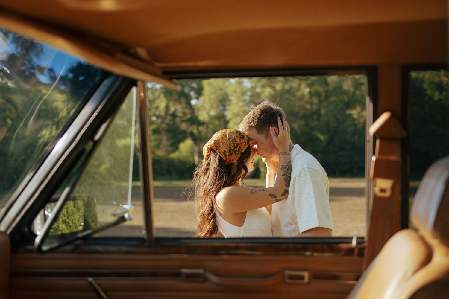 A couple shares a romantic moment kissing inside a vintage brown car during a golden hour outdoor photoshoot.