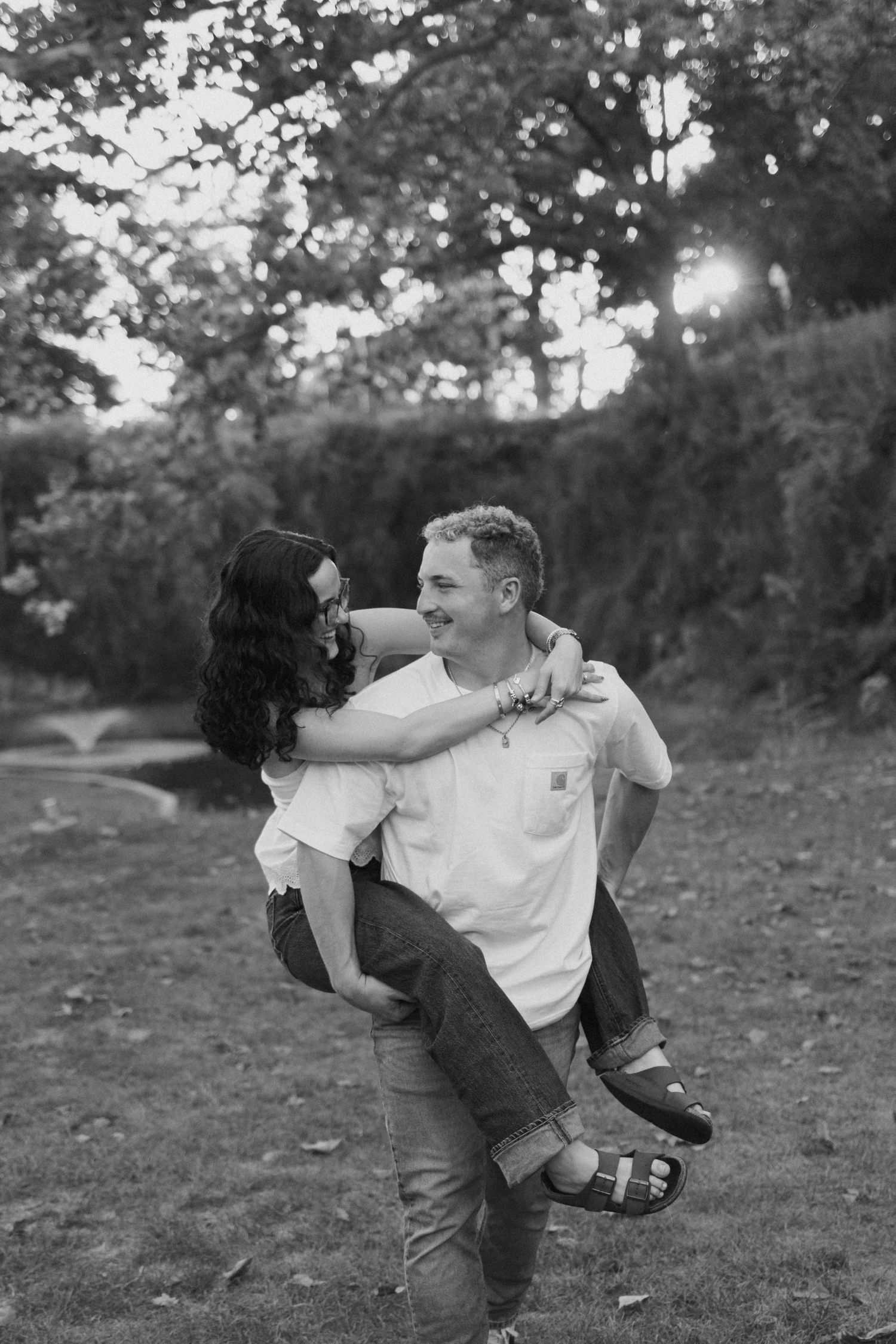 Black and white photo of a man carrying a woman piggyback in a park, both smiling joyfully.