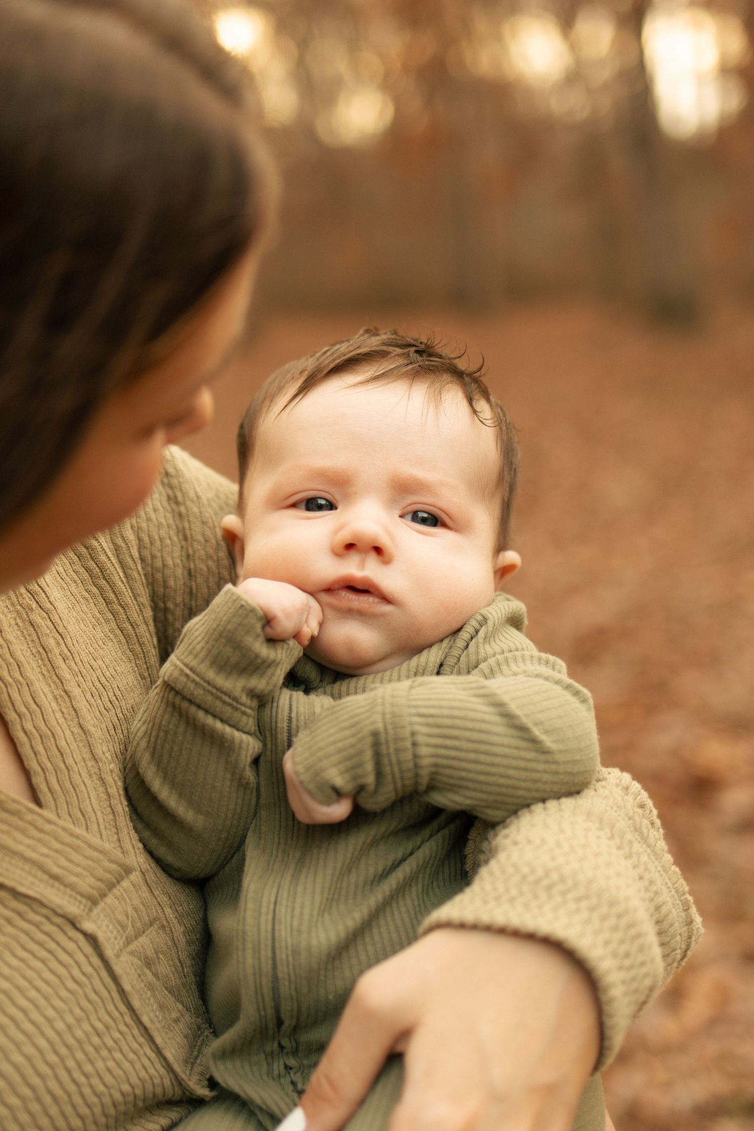 Close-up of alert newborn baby in green outfit being held by mother outdoors in fall setting.
