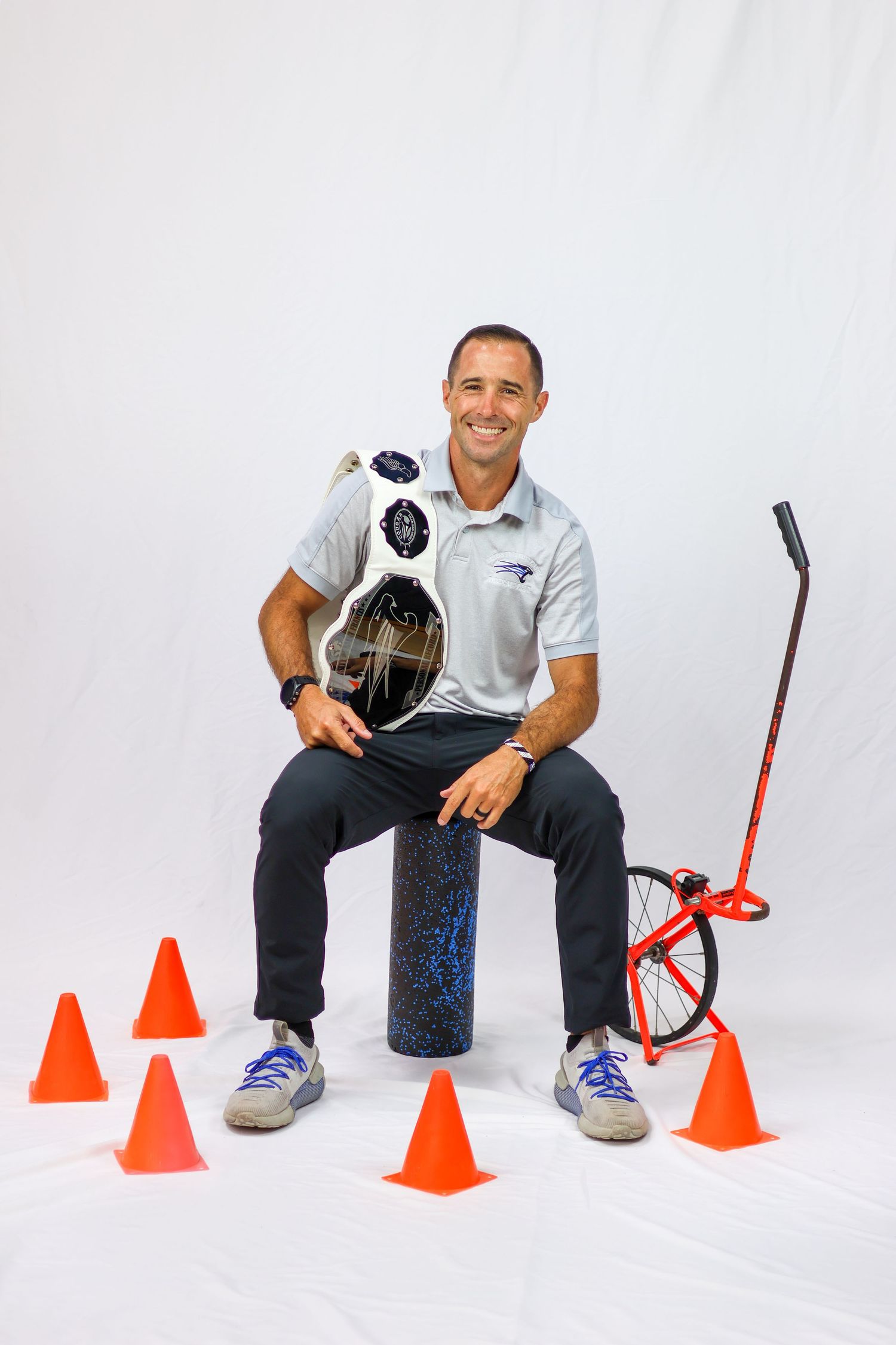 Athletic instructor sitting on exercise equipment surrounded by orange cones with fitness tools in a studio setting.
