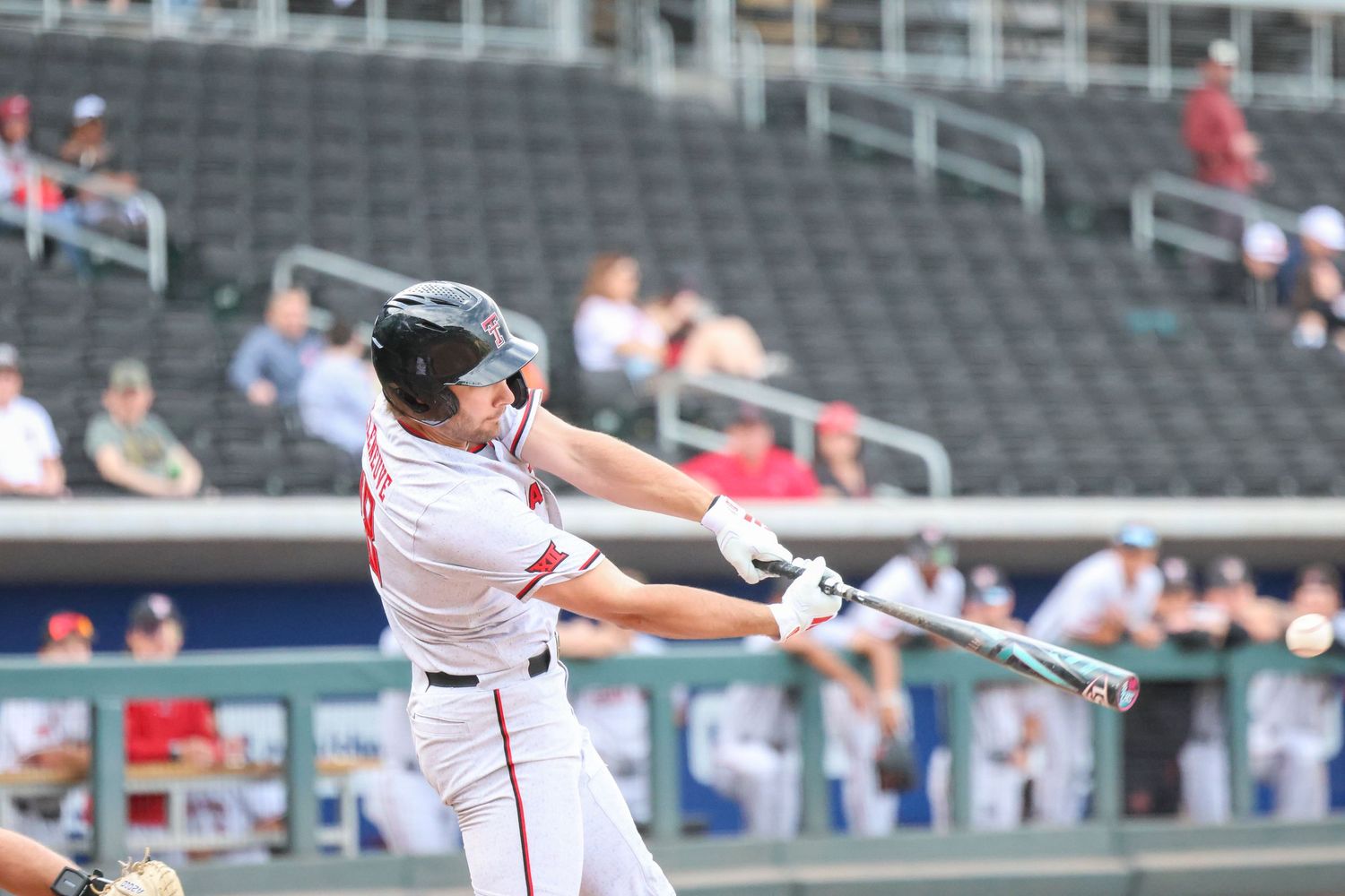 Baseball batter in white uniform swings bat during game with stadium seating visible in background.