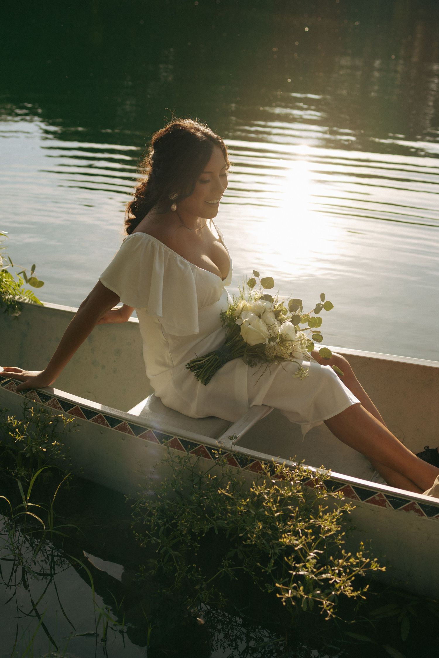 Bride in white off-shoulder dress holding bouquet of white roses, sitting in a canoe on a sunlit lake.