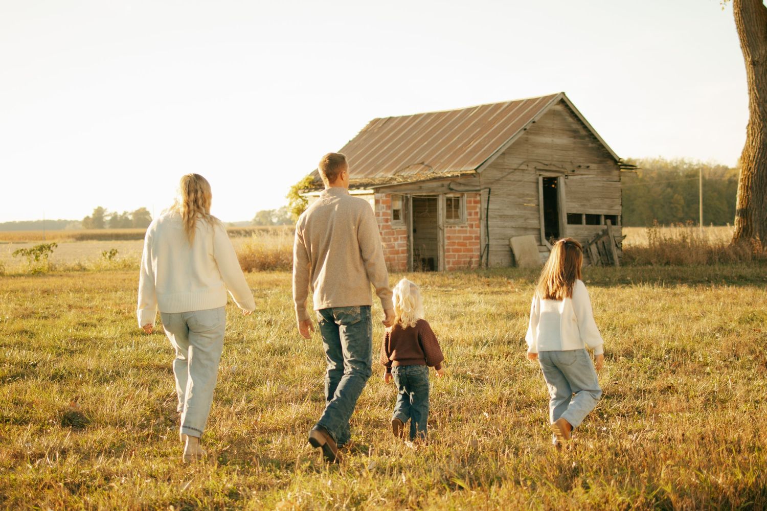 Family of four walking toward rustic farmhouse in golden field at sunset, parents and children holding hands.