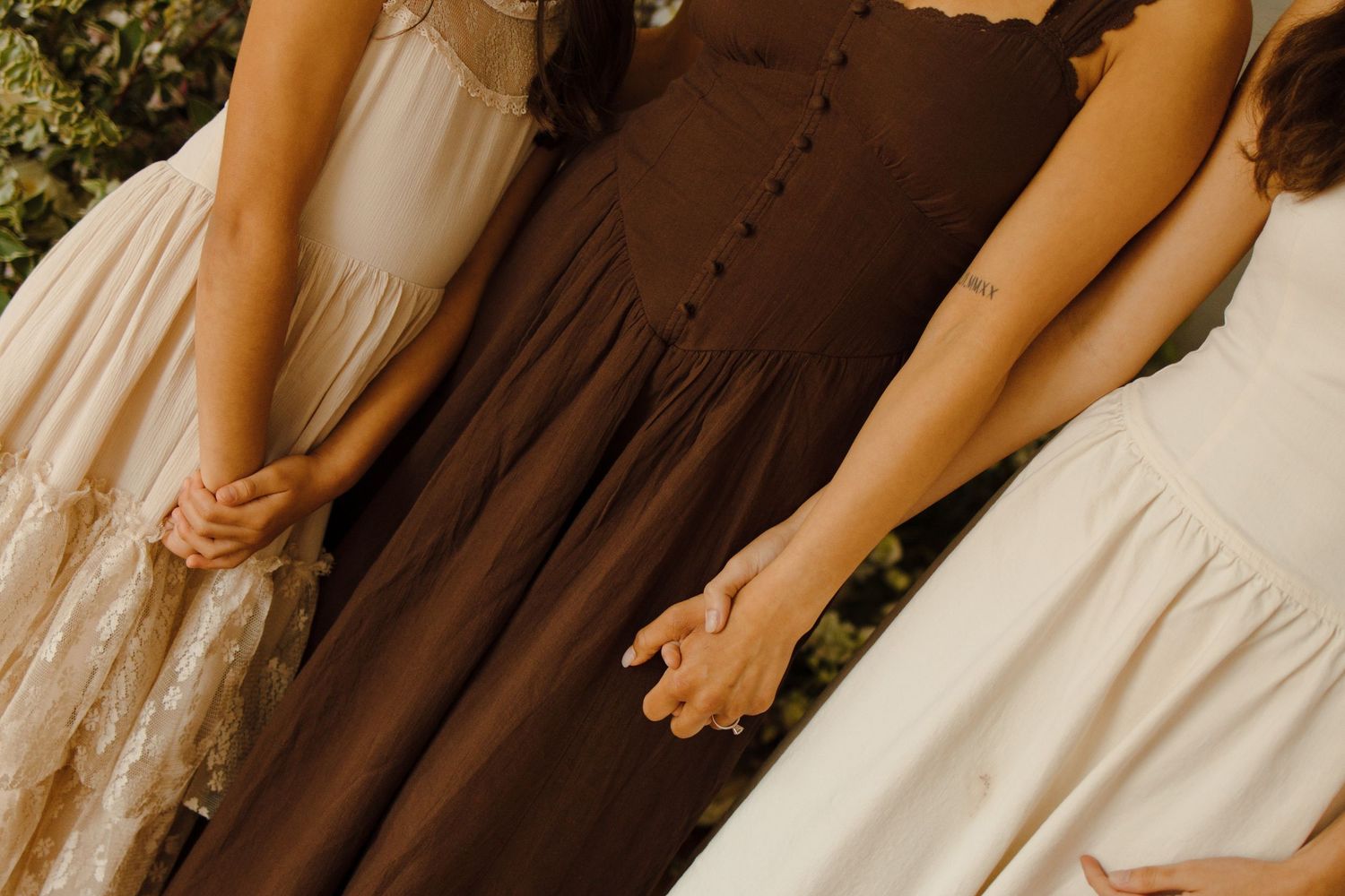 Three women in elegant dresses, brown, cream, and white, standing together holding hands at a wedding.