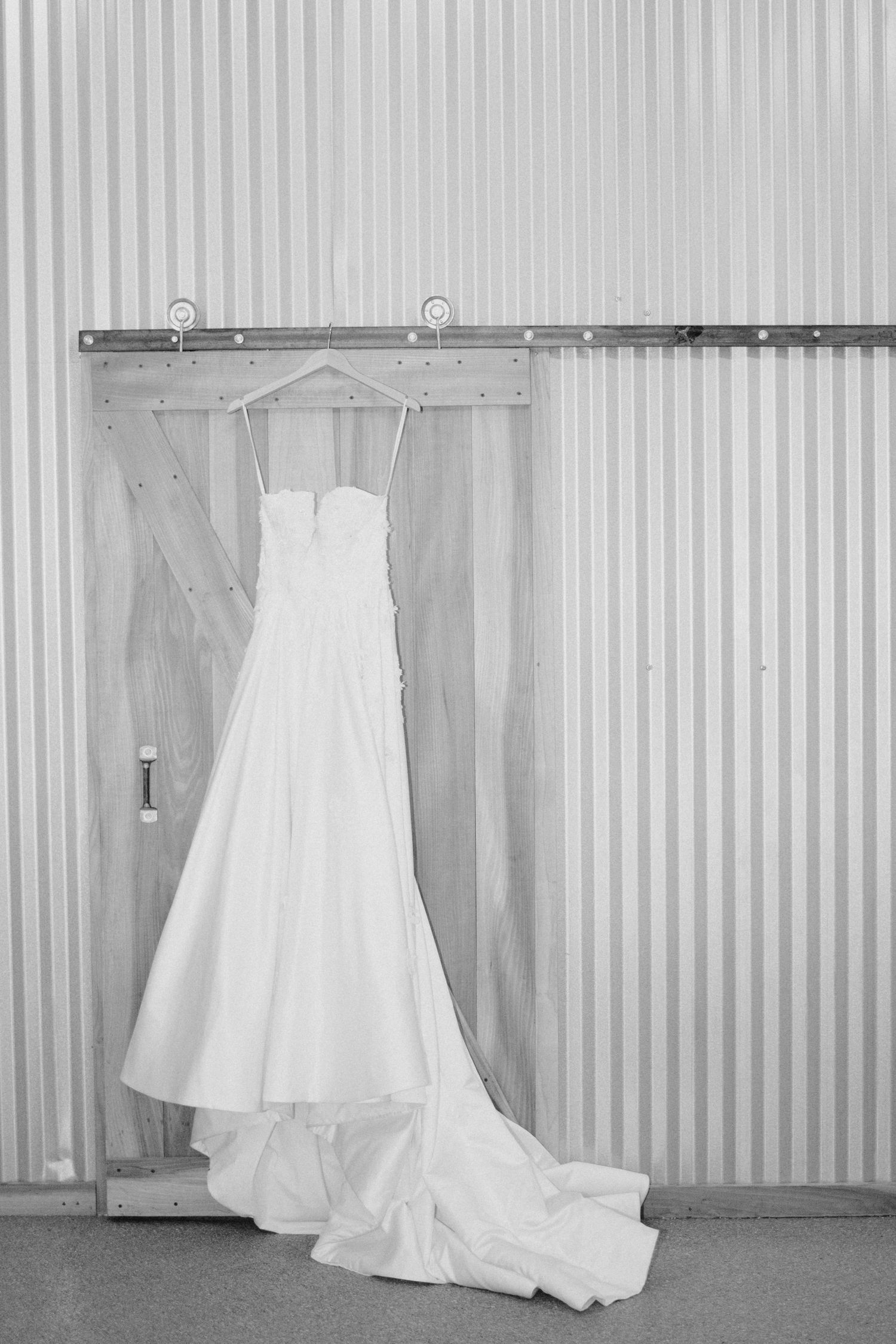 A white wedding gown with a long train hangs on a hanger against a corrugated metal wall in black and white.