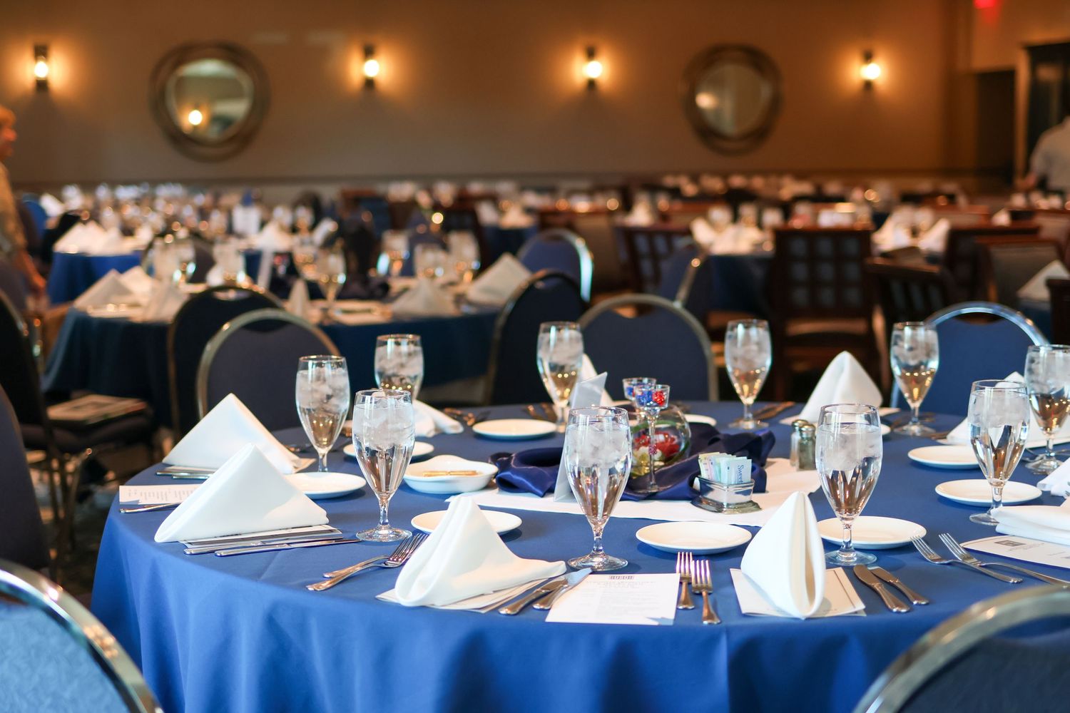 Elegant banquet hall with round tables draped in blue linens, set with white napkins and sparkling glassware.