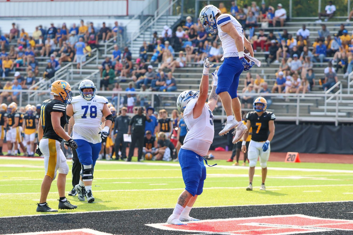 Football players celebrate a touchdown during a game with one player jumping high while another lifts him up.