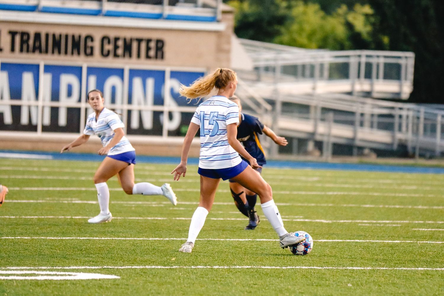 Soccer players compete for ball possession during match on turf field with training center visible in background.