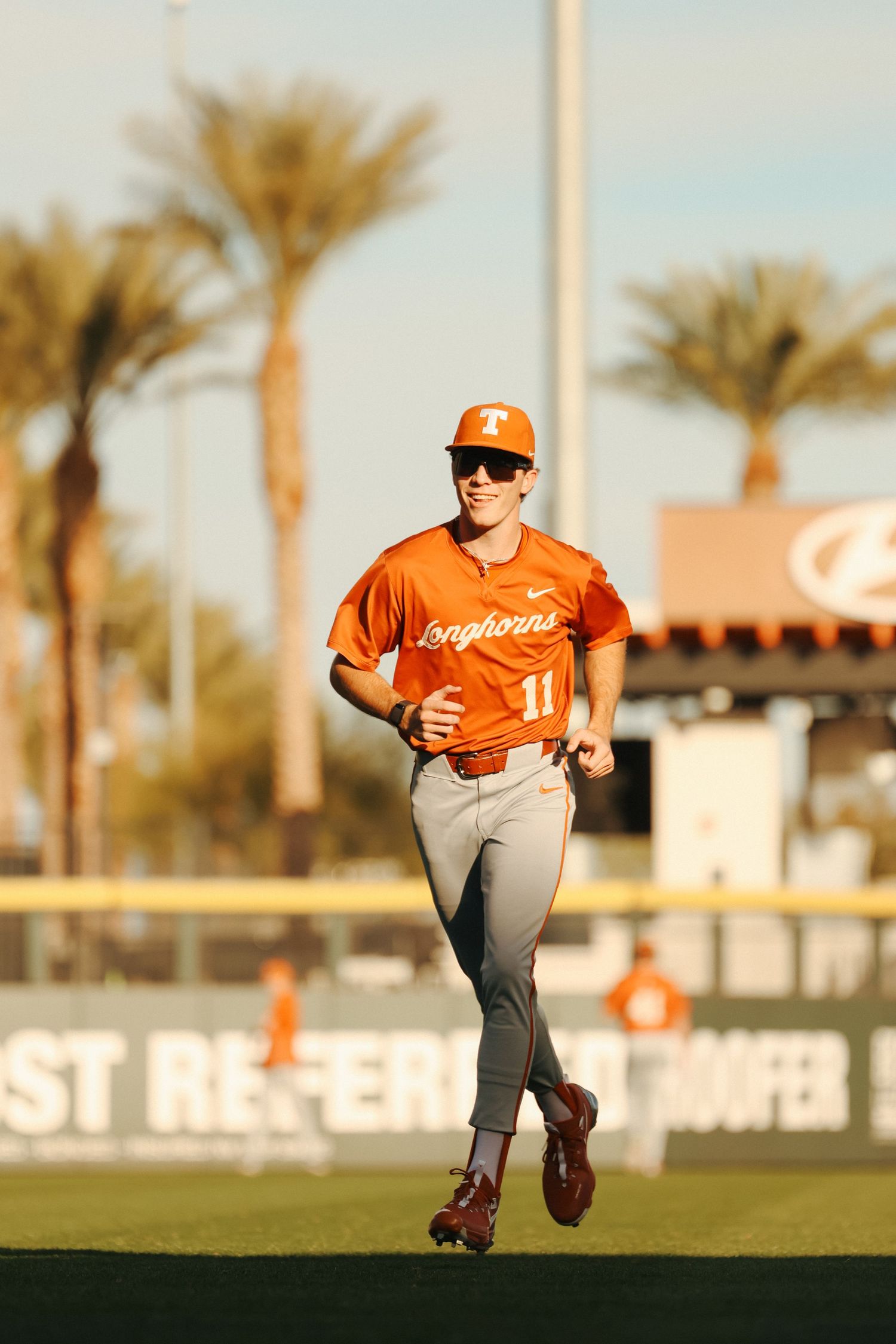 Baseball player in orange jersey running on field with palm trees in background during sunset practice session.