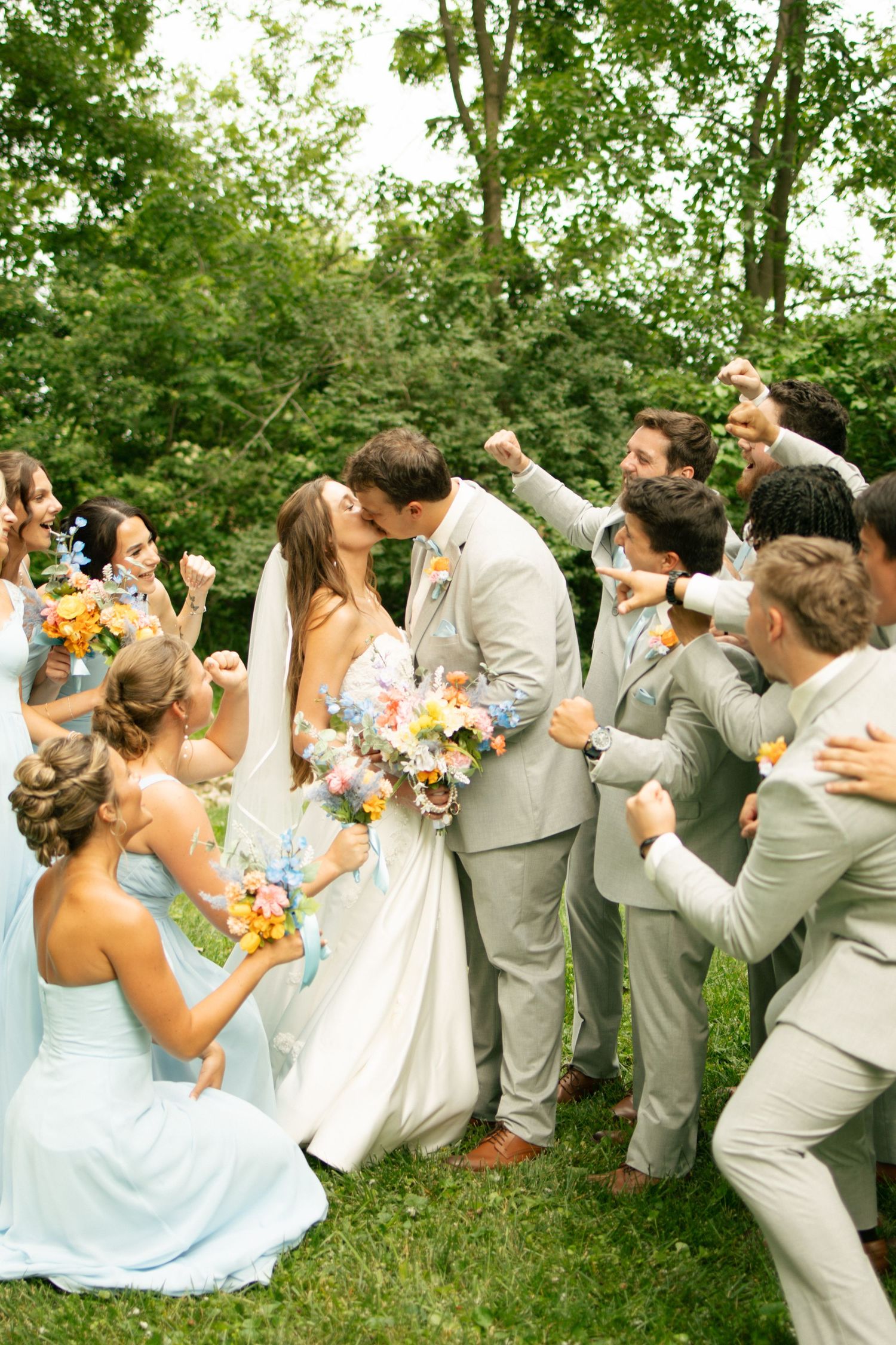 Bride and groom share a kiss surrounded by cheering wedding party in light blue and gray attire outdoors.
