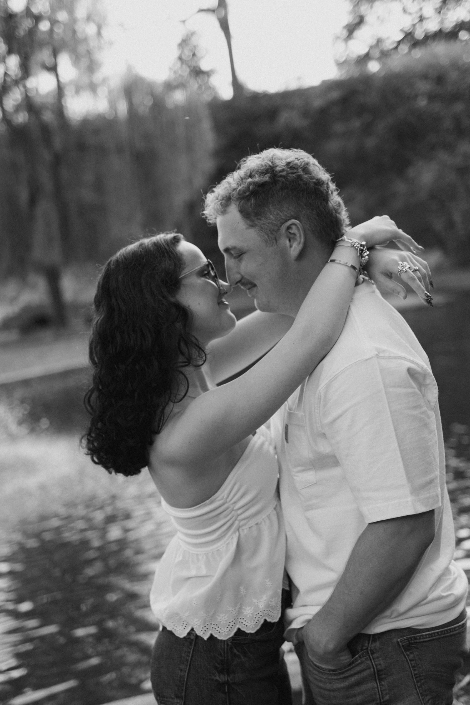 Black and white photo of a romantic couple embracing and about to kiss outdoors near a serene water backdrop.