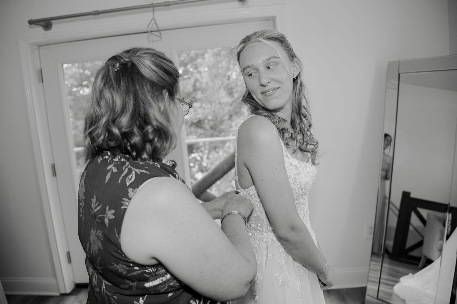 Black and white photo of a bride in a white dress being helped by a woman adjusting her gown before the wedding ceremony.