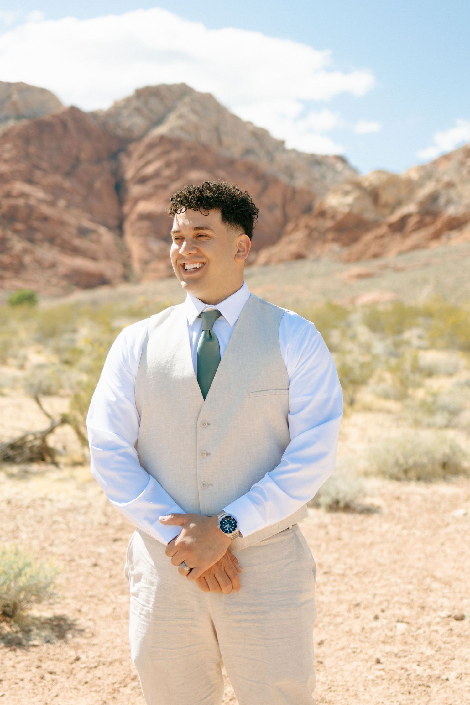 A groom in a light vest and tie smiles while standing in a desert setting with red rock formations.