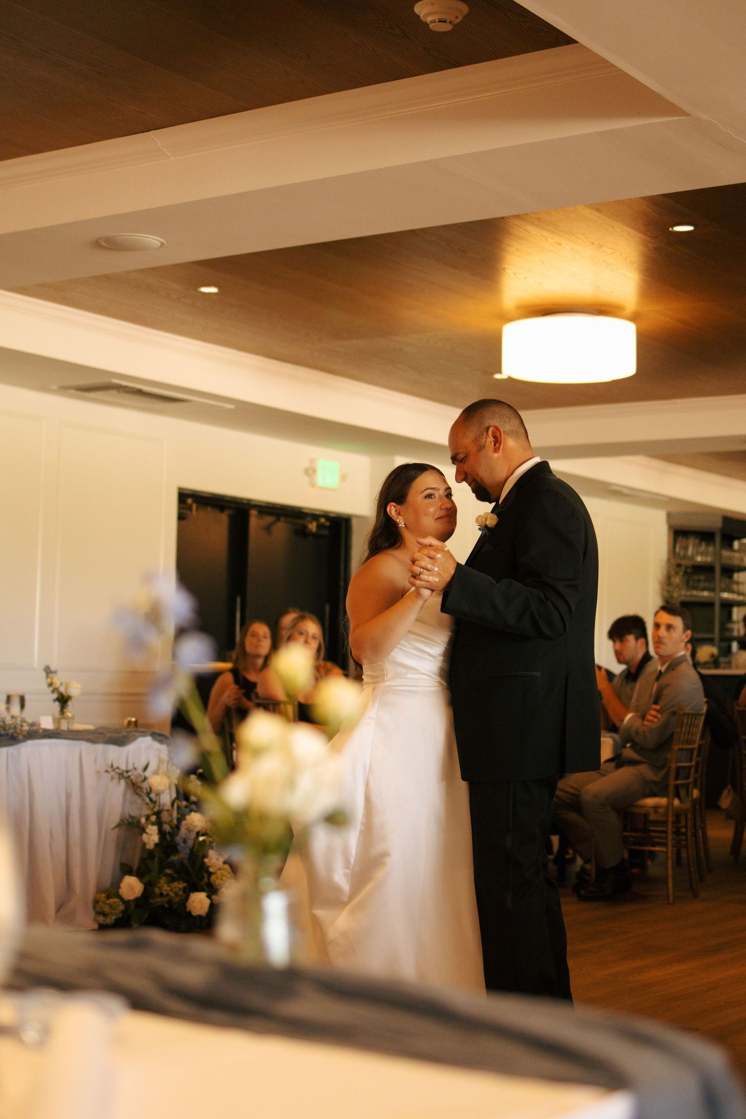 Bride and groom share their first dance at elegant wedding reception with floral arrangements and guests in background.