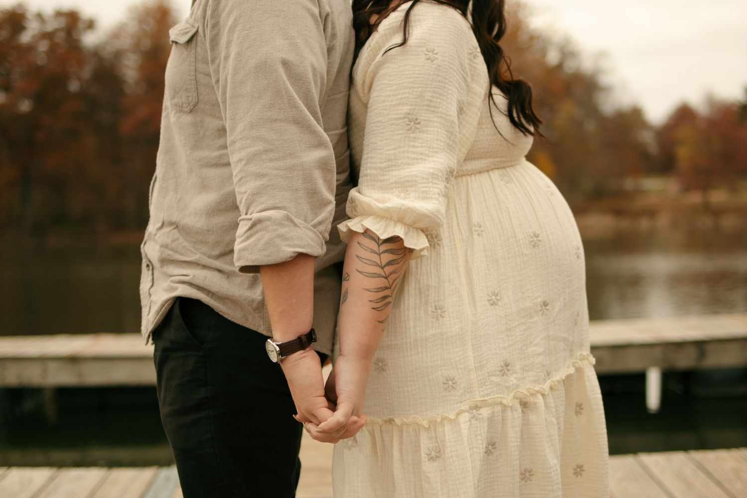 Pregnant couple holding hands on a wooden dock near a lake surrounded by autumn trees.
