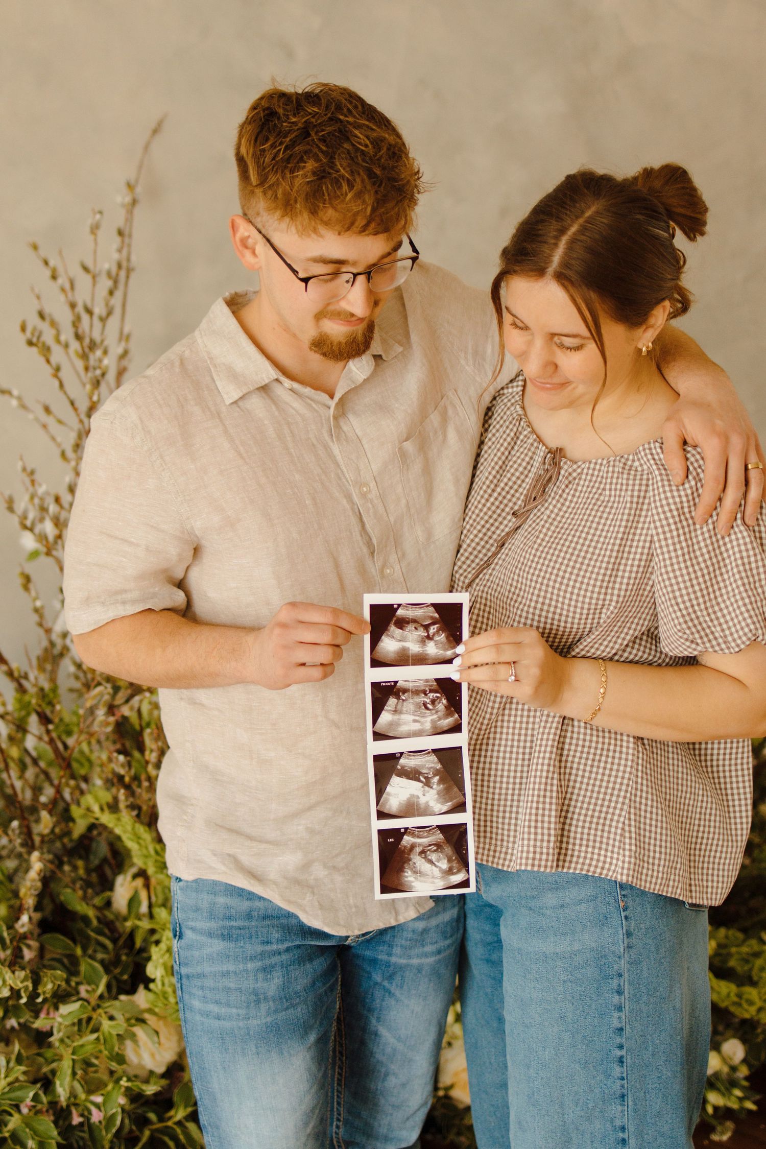 Couple smiling and holding ultrasound photos, announcing pregnancy while standing in front of green plants.