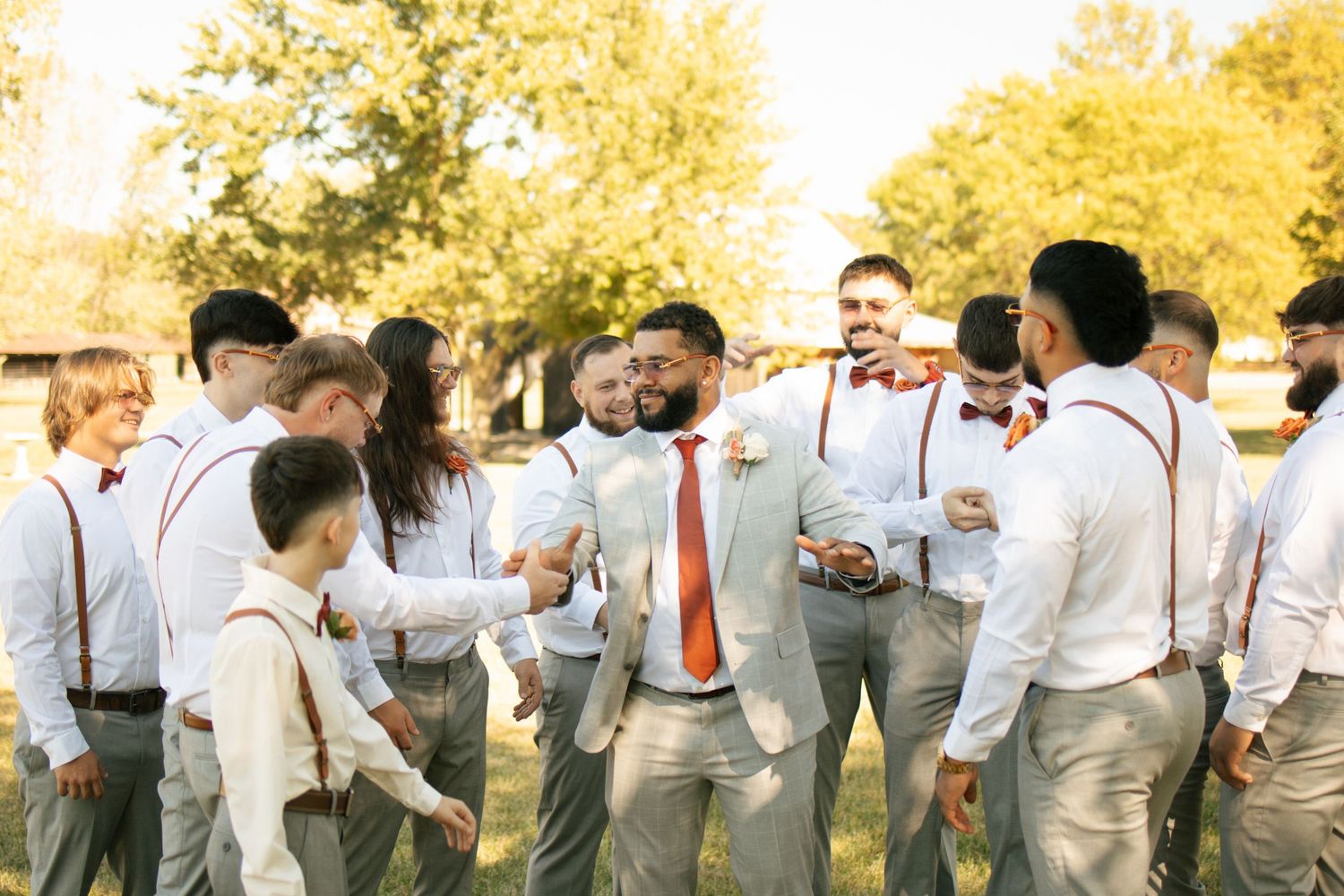 Groomsmen in white shirts with rust bow ties and suspenders laughing together outdoors at a wedding celebration.